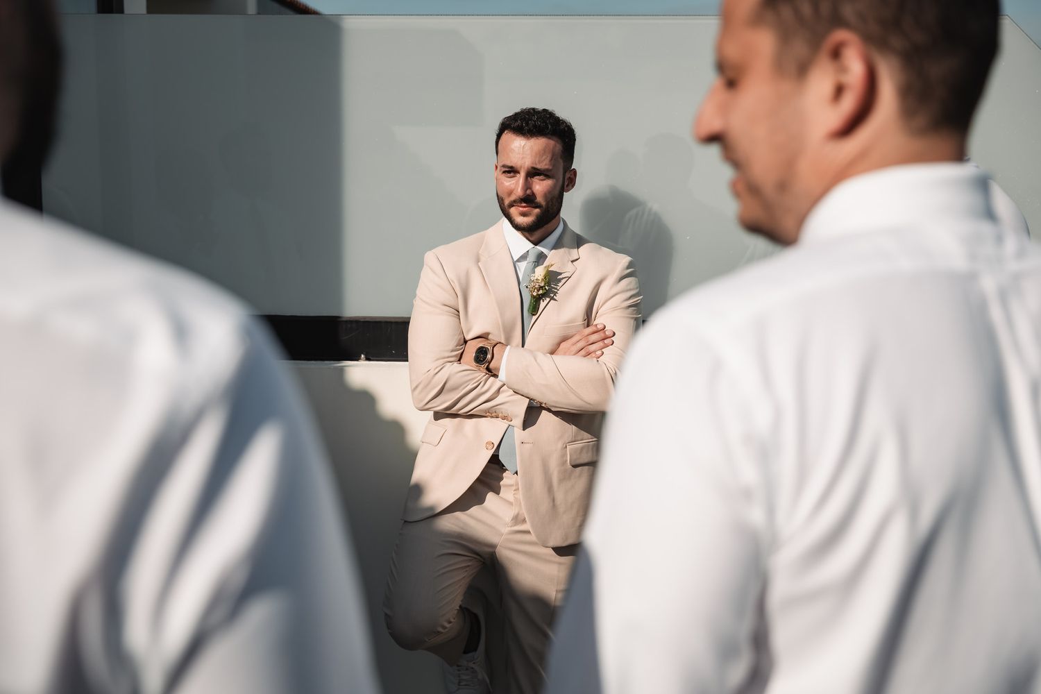 Groom in beige suit sits with arms crossed smiling at groomsmen during wedding preparations.