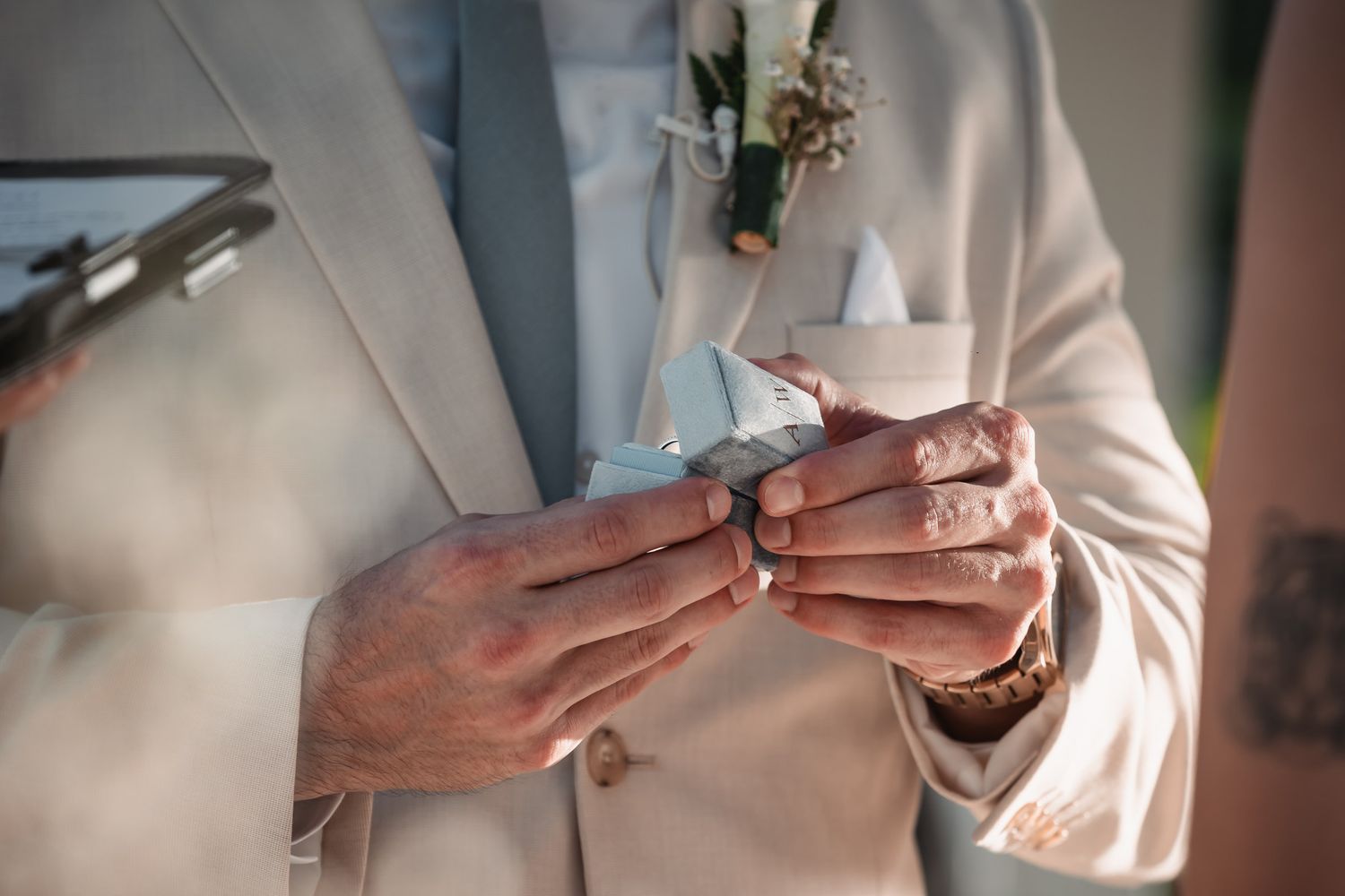 Groom in white suit reads wedding vows from paper with boutonniere at outdoor ceremony.