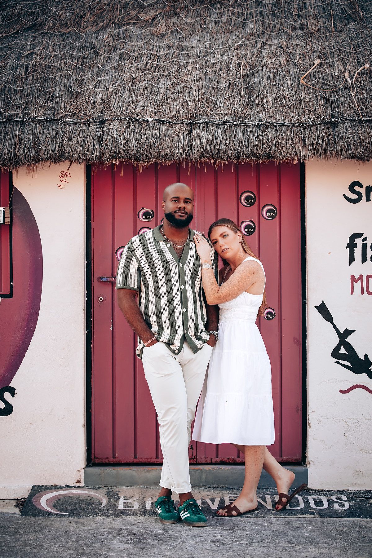 Stylish interracial couple posing together in front of a rustic thatched-roof building with red doors.
