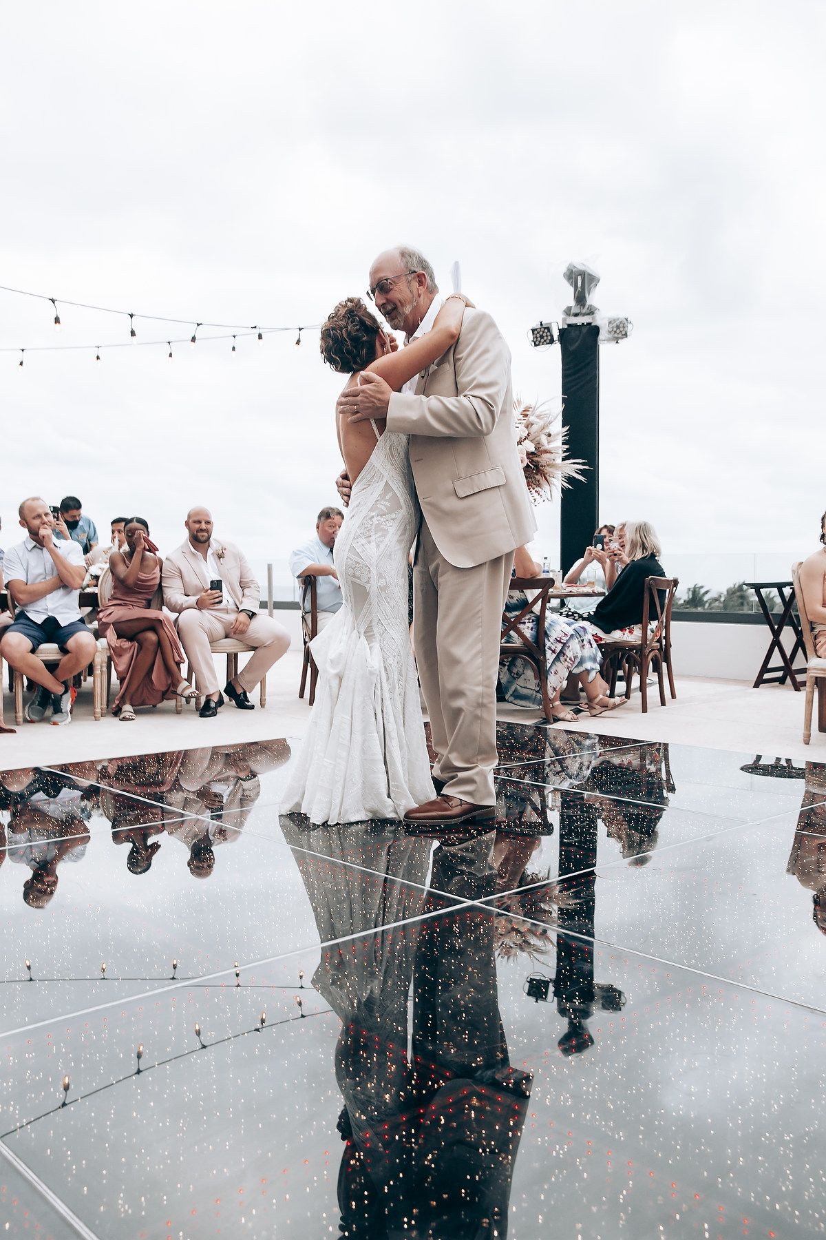 Couple dancing on reflective surface at outdoor wedding reception with string lights.