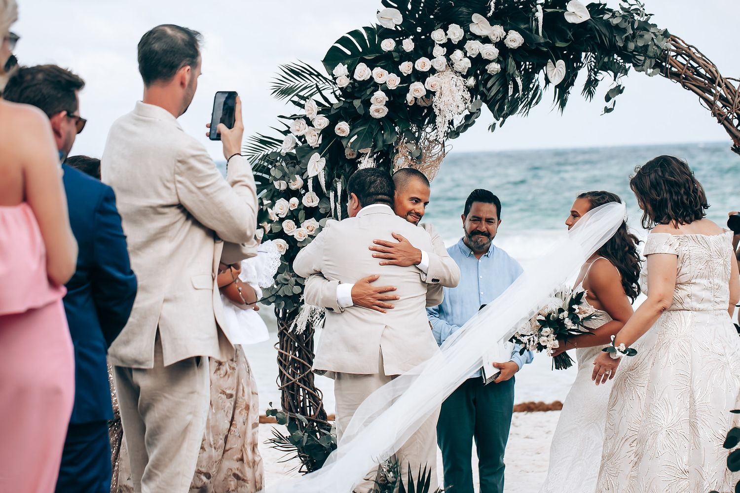 Wedding guests gather on tropical beach to celebrate ceremony under palm trees.