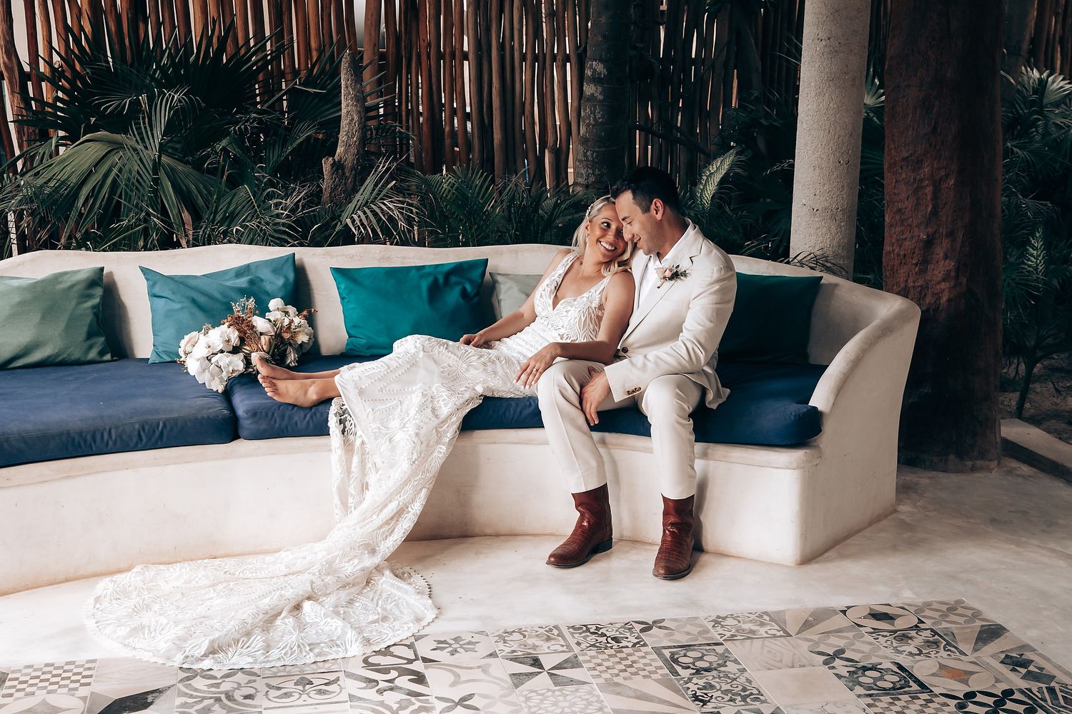 A romantic wedding couple relaxes on a blue couch with tropical decor and palm trees in the background.