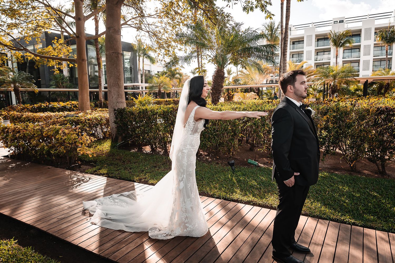 Bride in white gown taps groom's shoulder for first look on wooden deck surrounded by tropical gardens.