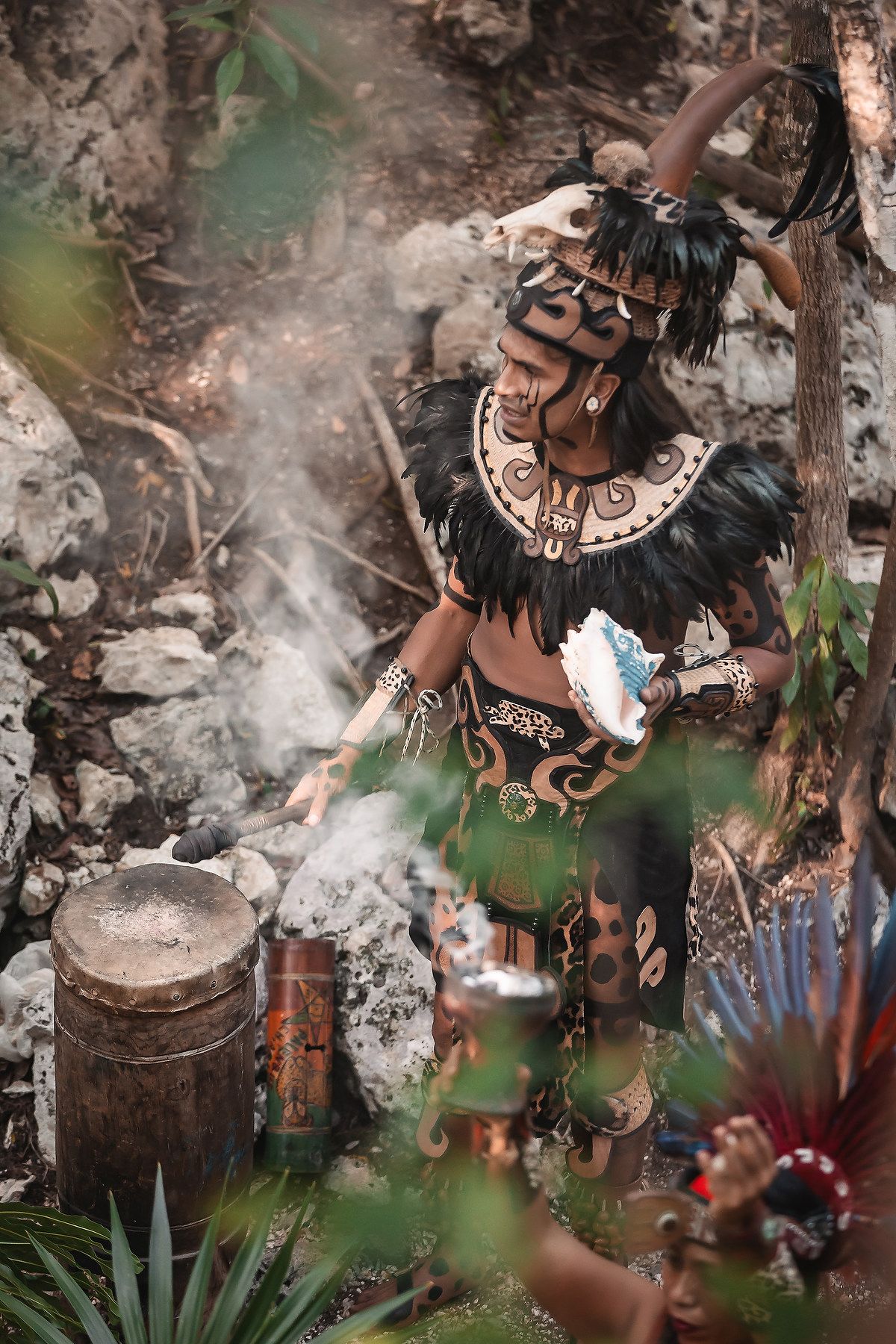 Mayan warrior in traditional costume with feathers, skull headdress, and body paint performs ritual ceremony with smoke.