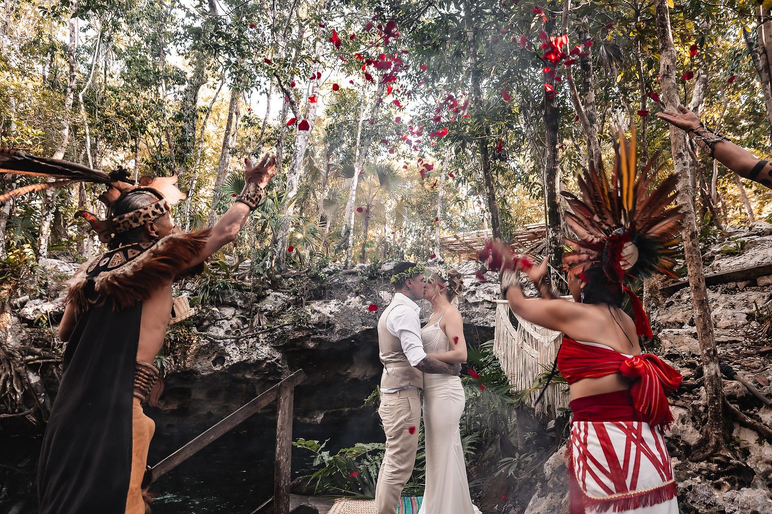 Couple kissing during Mayan wedding ceremony in jungle cenote with colorful headdressed officiants tossing flower petals.