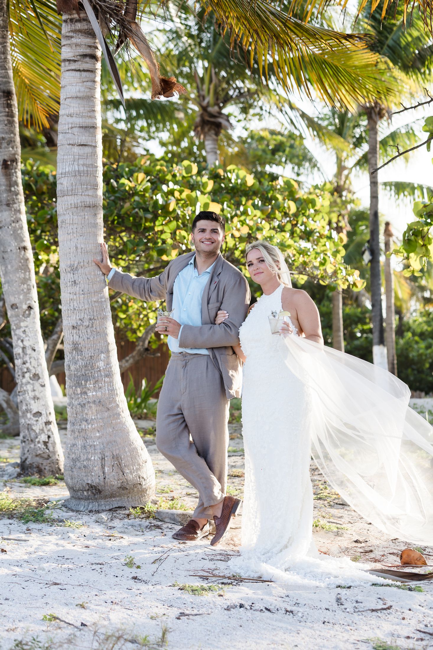 Bride and groom pose among tropical palm trees on sandy beach, bride's white gown flowing in the breeze.