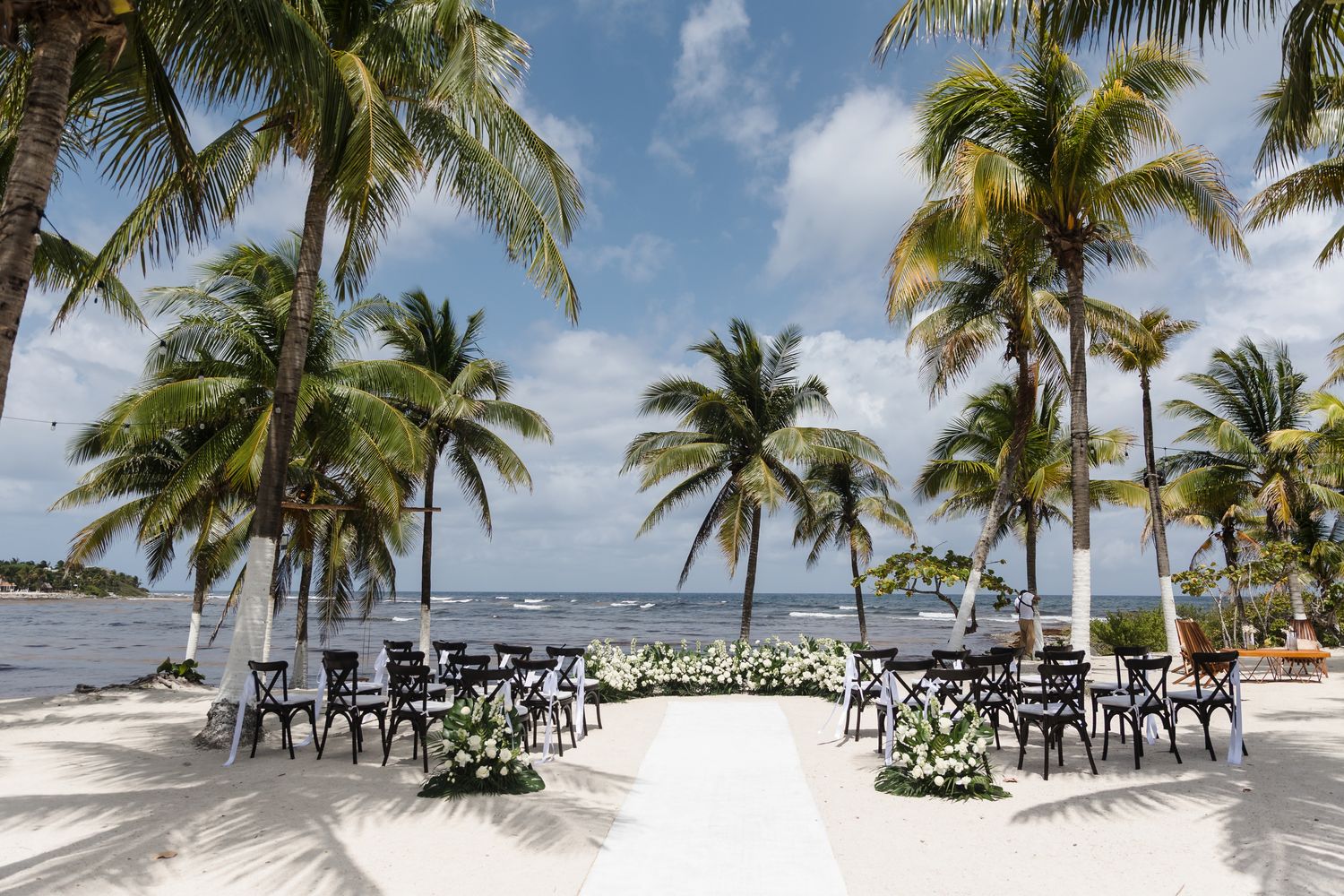 Elegant Playa del Carmen beach wedding setup with a white floral aisle, dark chairs, palm trees, and ocean views.
