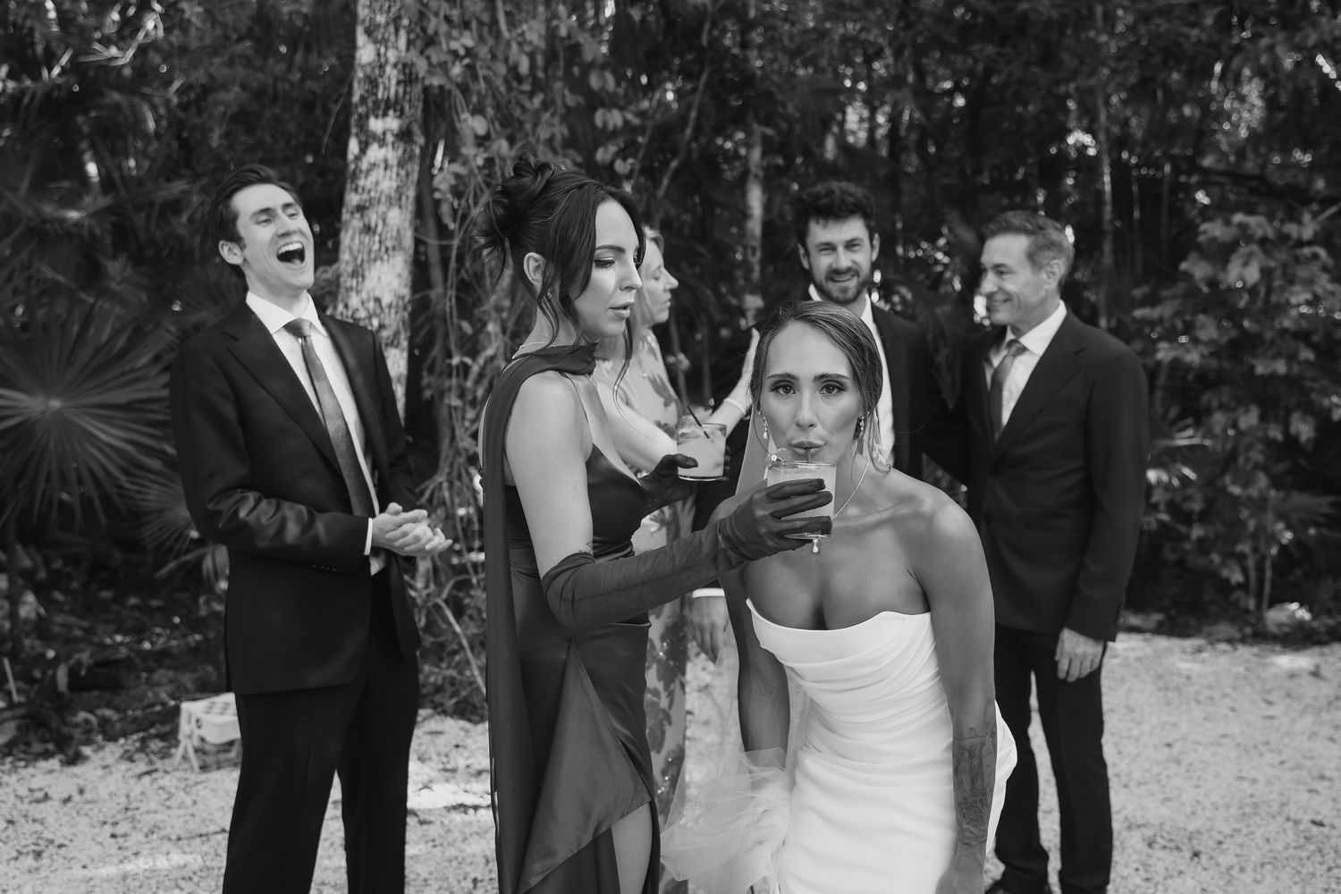 Black and white photo of bride drinking champagne with wedding guests laughing in a forest setting.