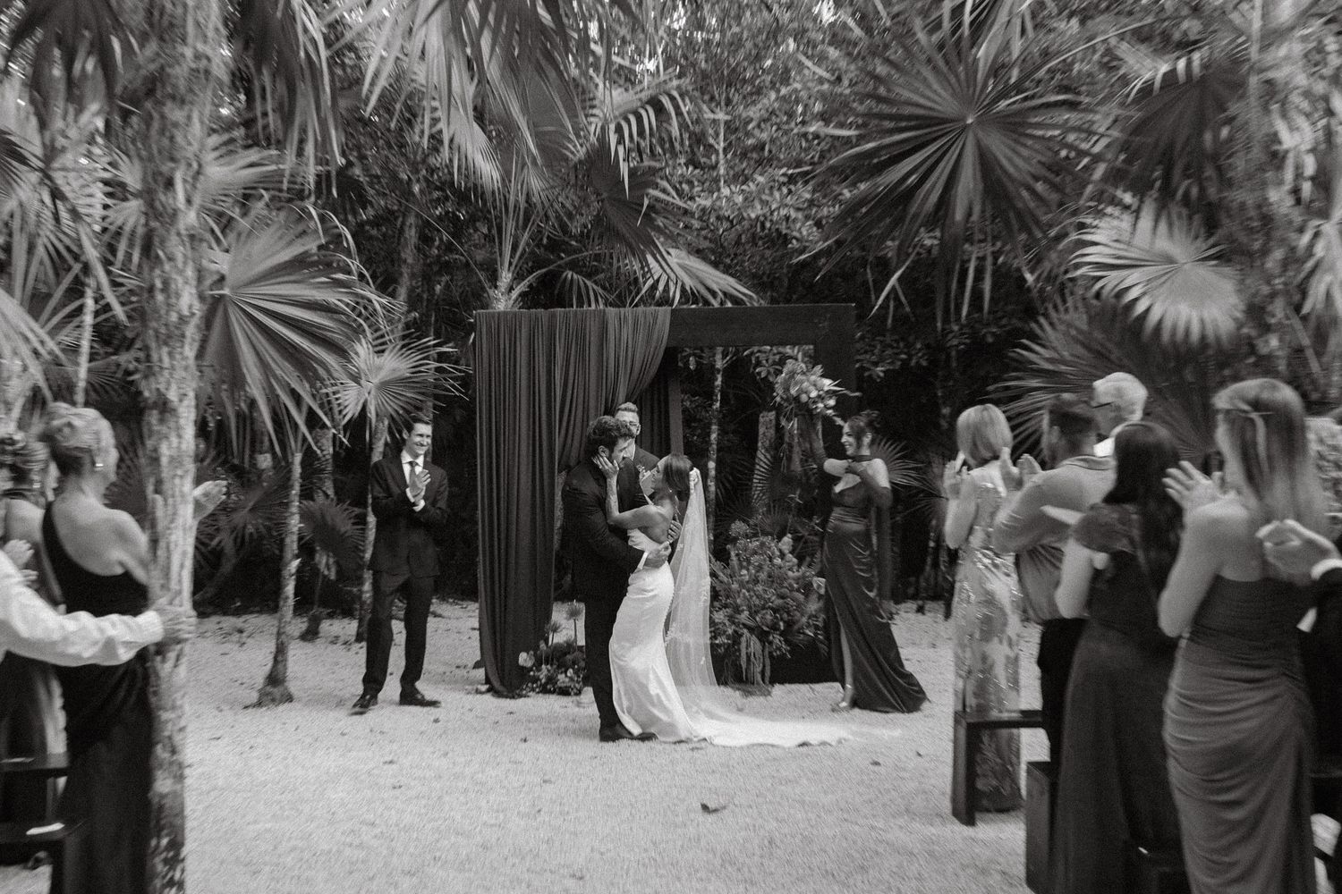 Black and white photo of couple sharing first kiss at outdoor tropical wedding ceremony.