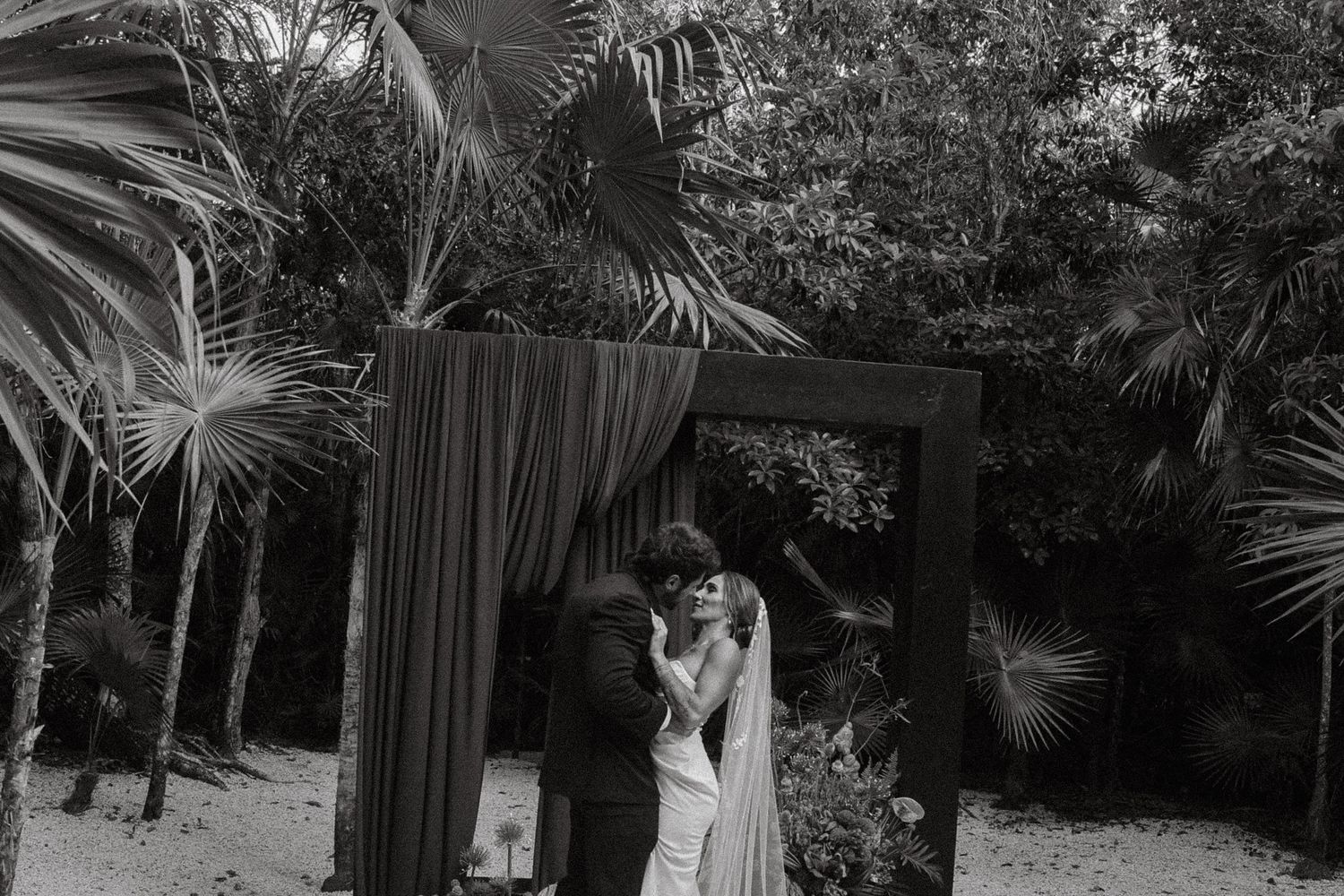 Black and white wedding photo of bride and groom kissing under a floral arch surrounded by tropical palm trees.