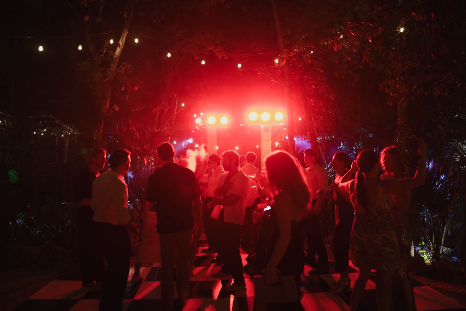 Crowd of people dancing outdoors at night under red stage lights and trees at an evening party event.