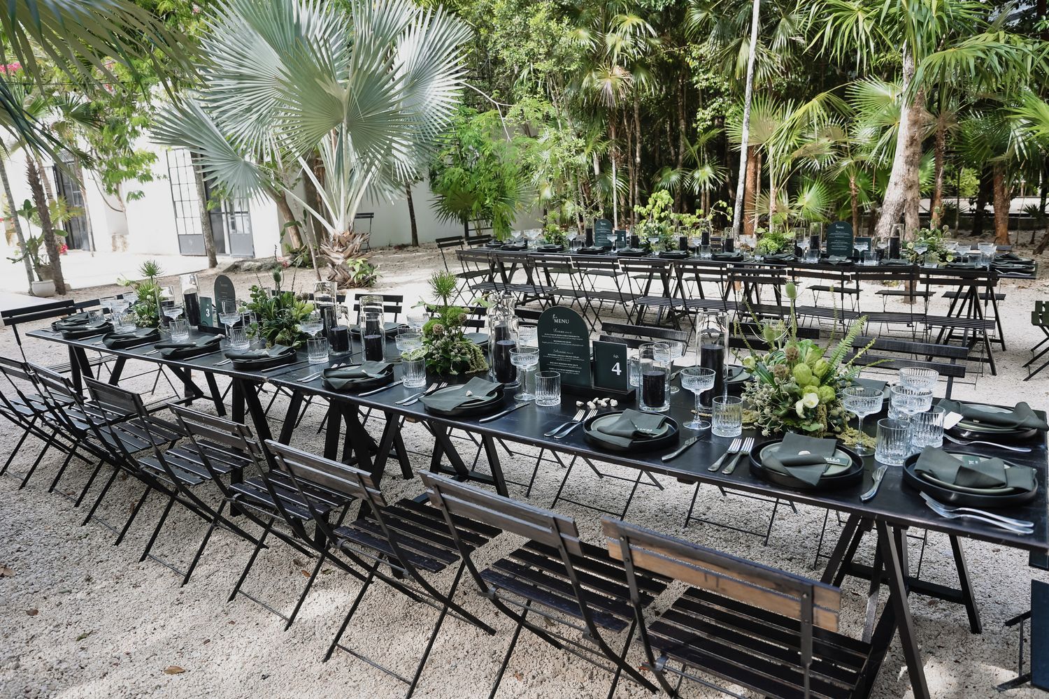 Elegant outdoor dining setup with black tables, chairs, and dark plates amid lush tropical palm trees.