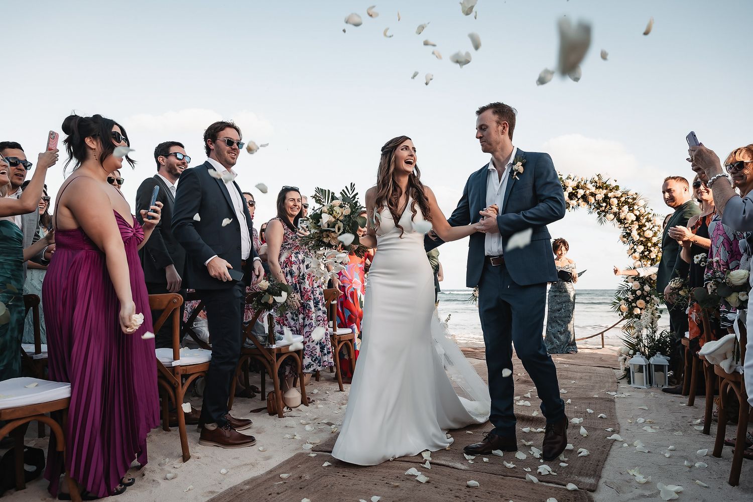 Bride and groom walk down the aisle as guests toss flower petals at an outdoor beach wedding ceremony.