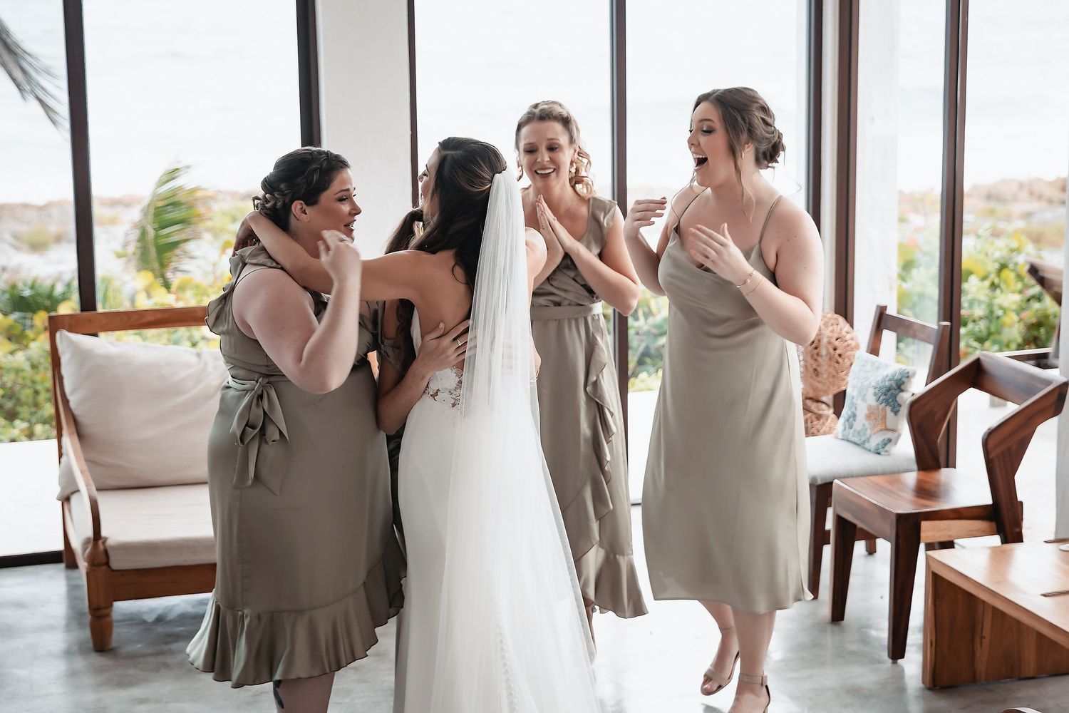 Bridesmaids in sage green dresses joyfully reacting to the bride in a bright tropical wedding venue.