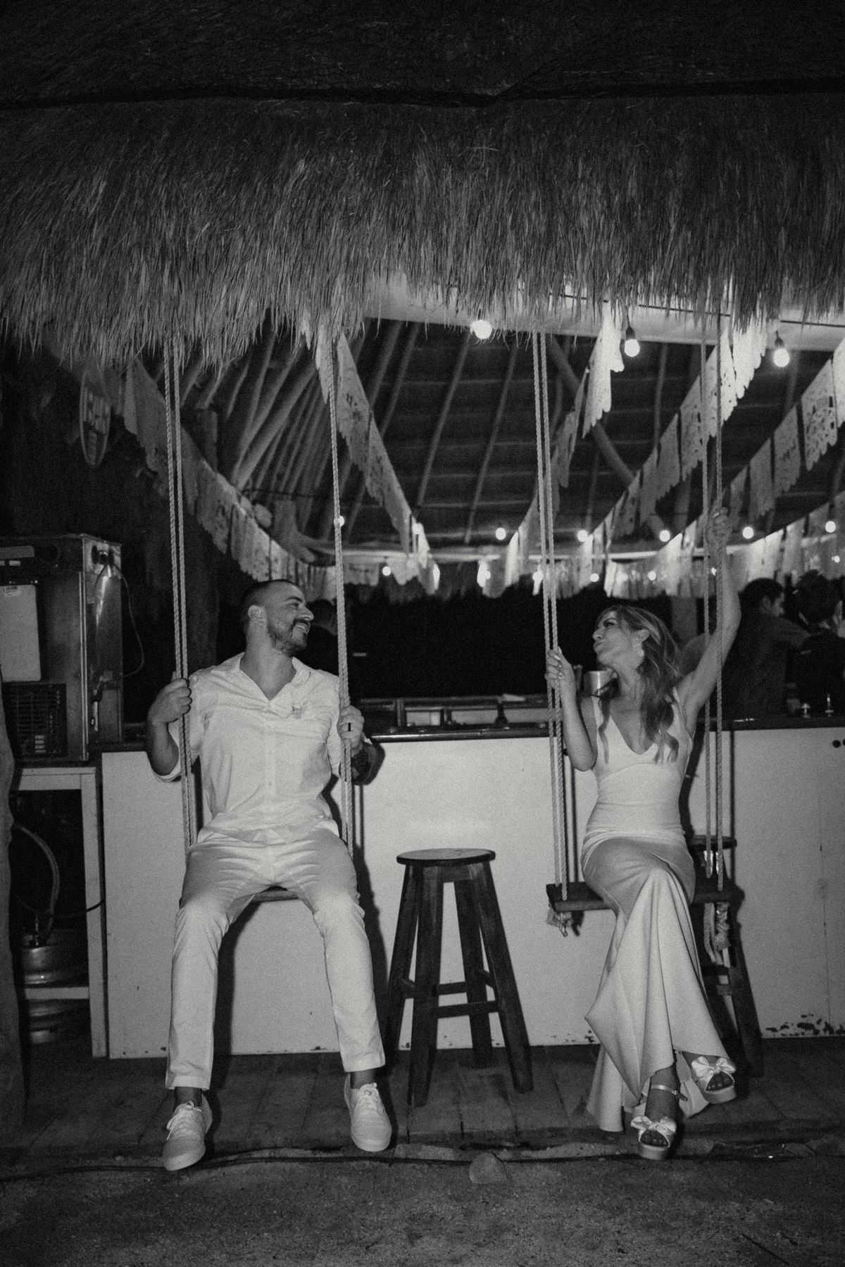 Bride and groom sit on swings laughing at a thatched roof beach bar at night.