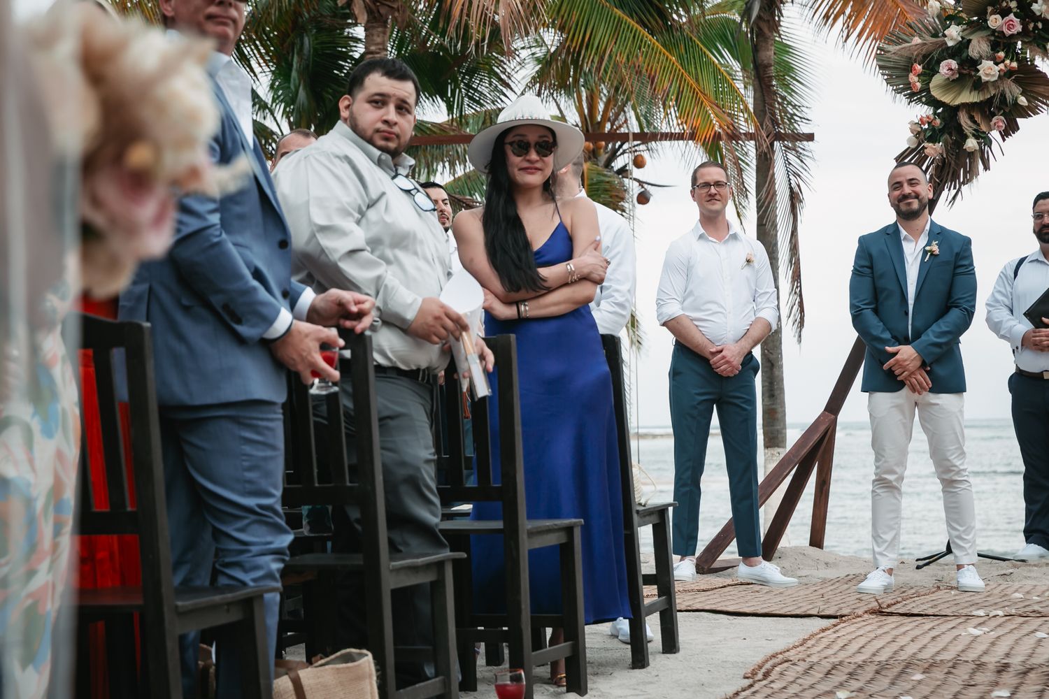 Wedding guests in elegant attire stand and watch a beach ceremony near palm trees and floral decorations.