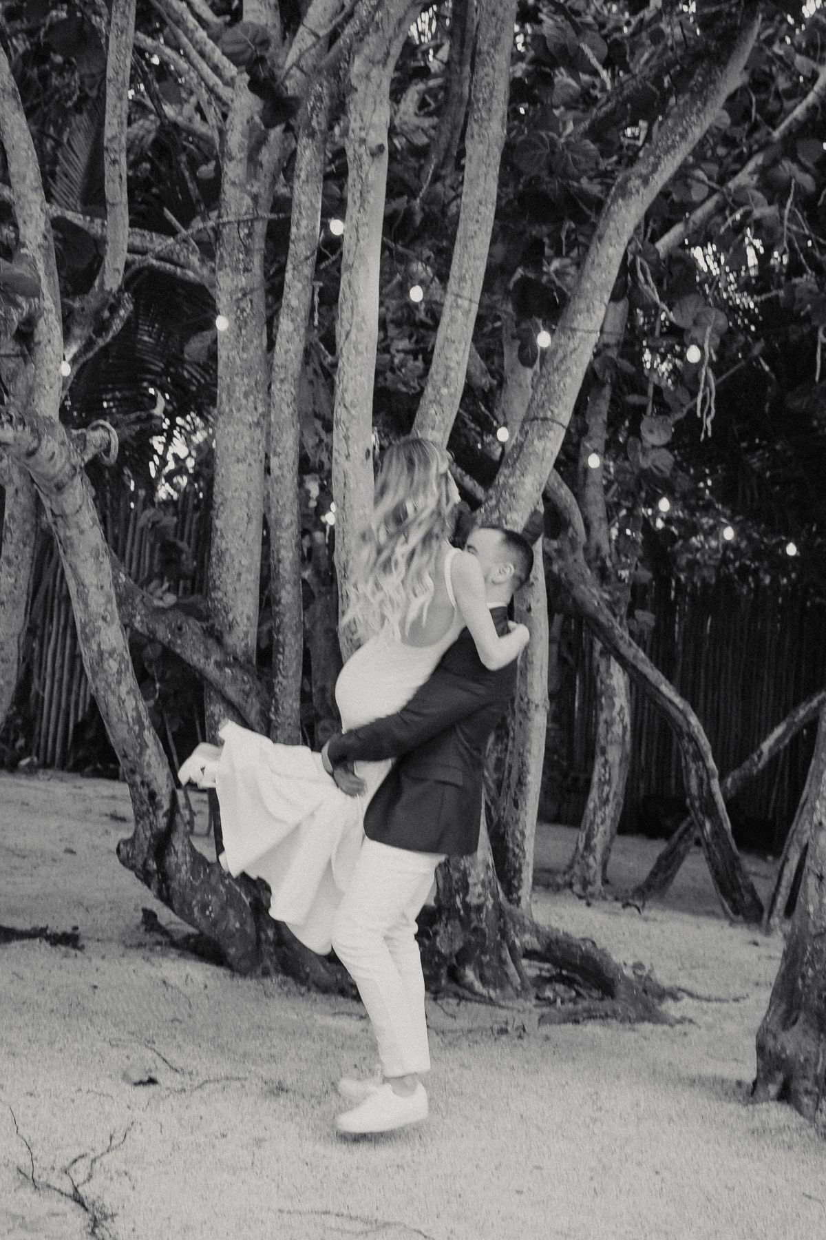 Black and white romantic photo of groom lifting bride among trees decorated with string lights on beach.