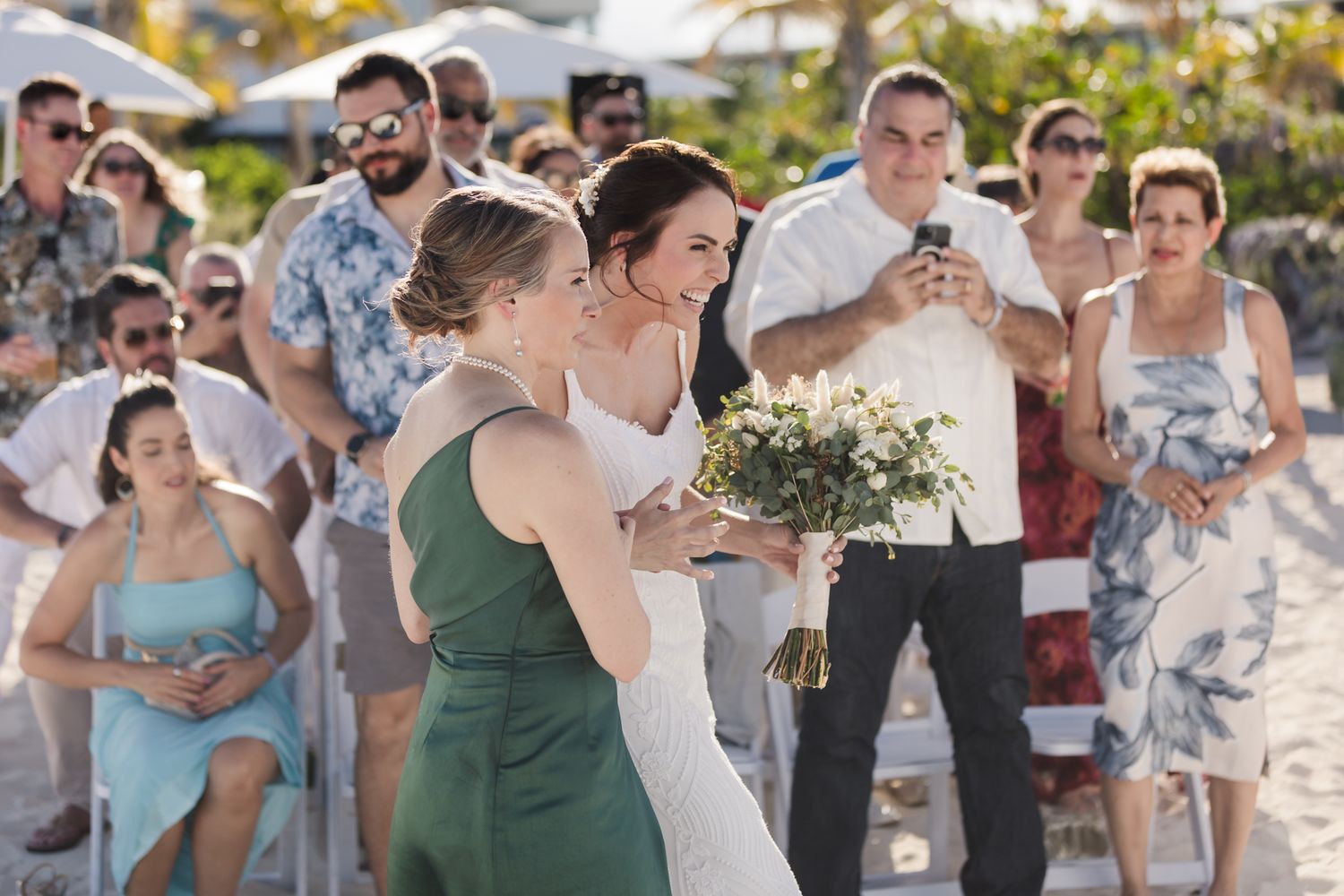 Bride in white gown walks with bridesmaid in green dress holding bouquet at outdoor beach wedding ceremony.