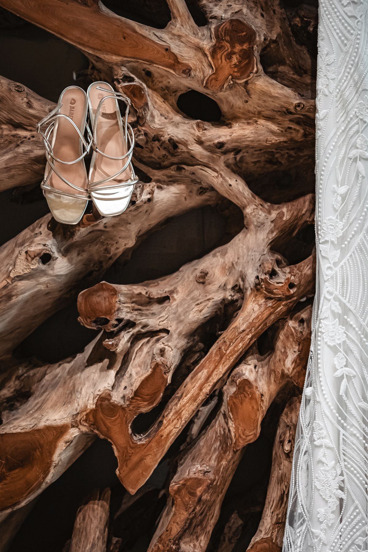 Elegant strappy bridal heels displayed on rustic driftwood sculpture with intricate natural curves and warm brown tones.
