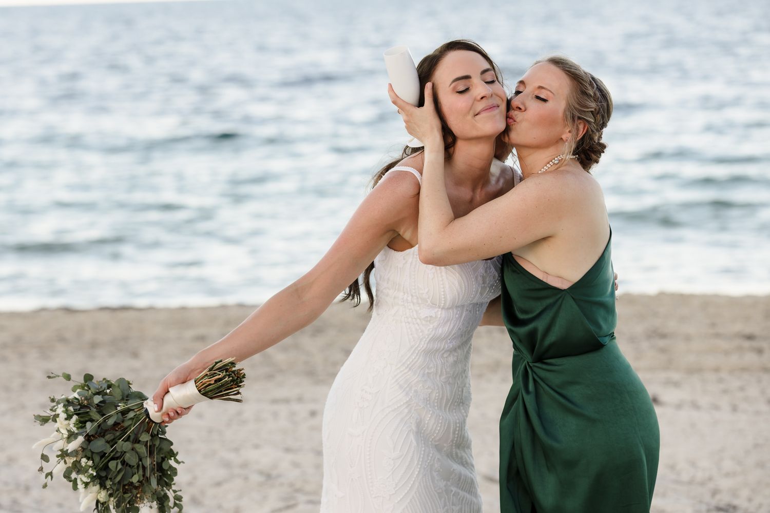 Bride in white dress holding greenery bouquet receives kiss on cheek from woman in green gown on sandy beach.