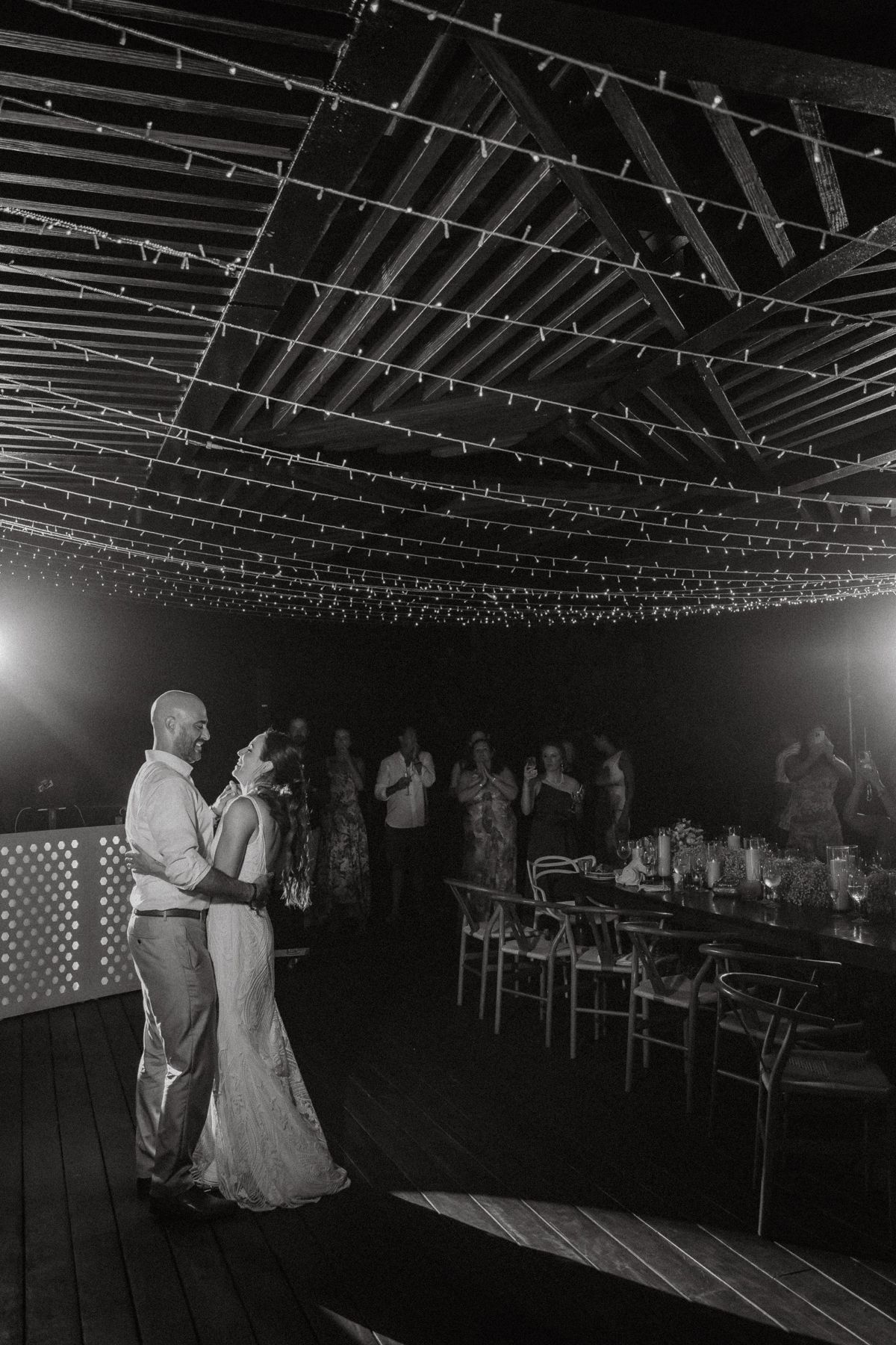 Couple shares first dance under string lights in rustic barn venue, guests watching in black and white wedding photo.