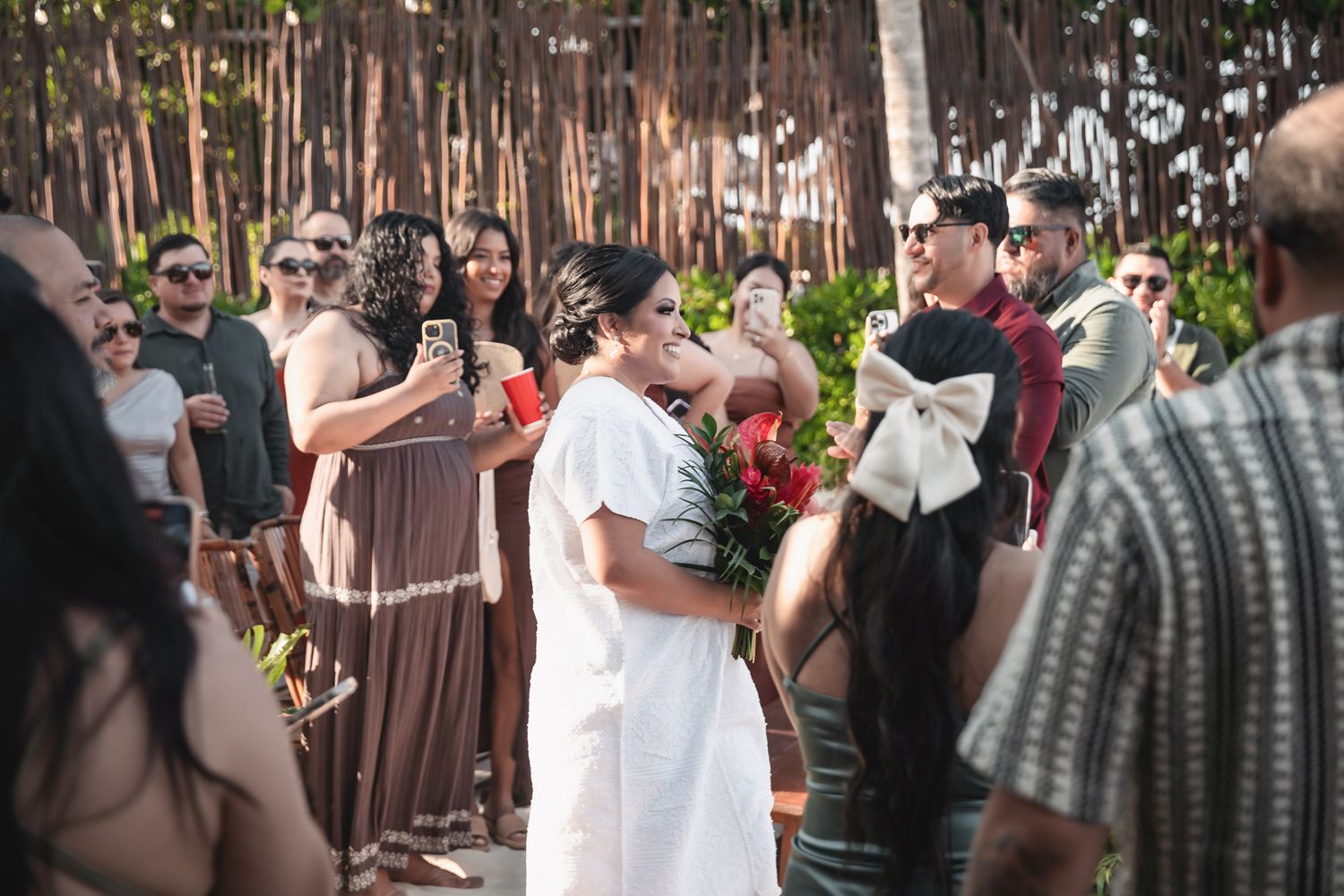 Bride in white dress holds red bouquet surrounded by guests photographing at outdoor wedding ceremony.