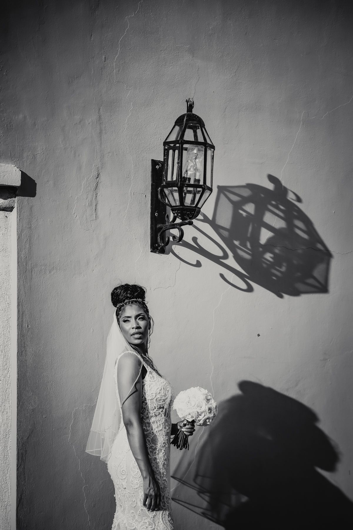 Elegant bride in lace gown holds bouquet beside vintage wall lantern in black and white portrait.