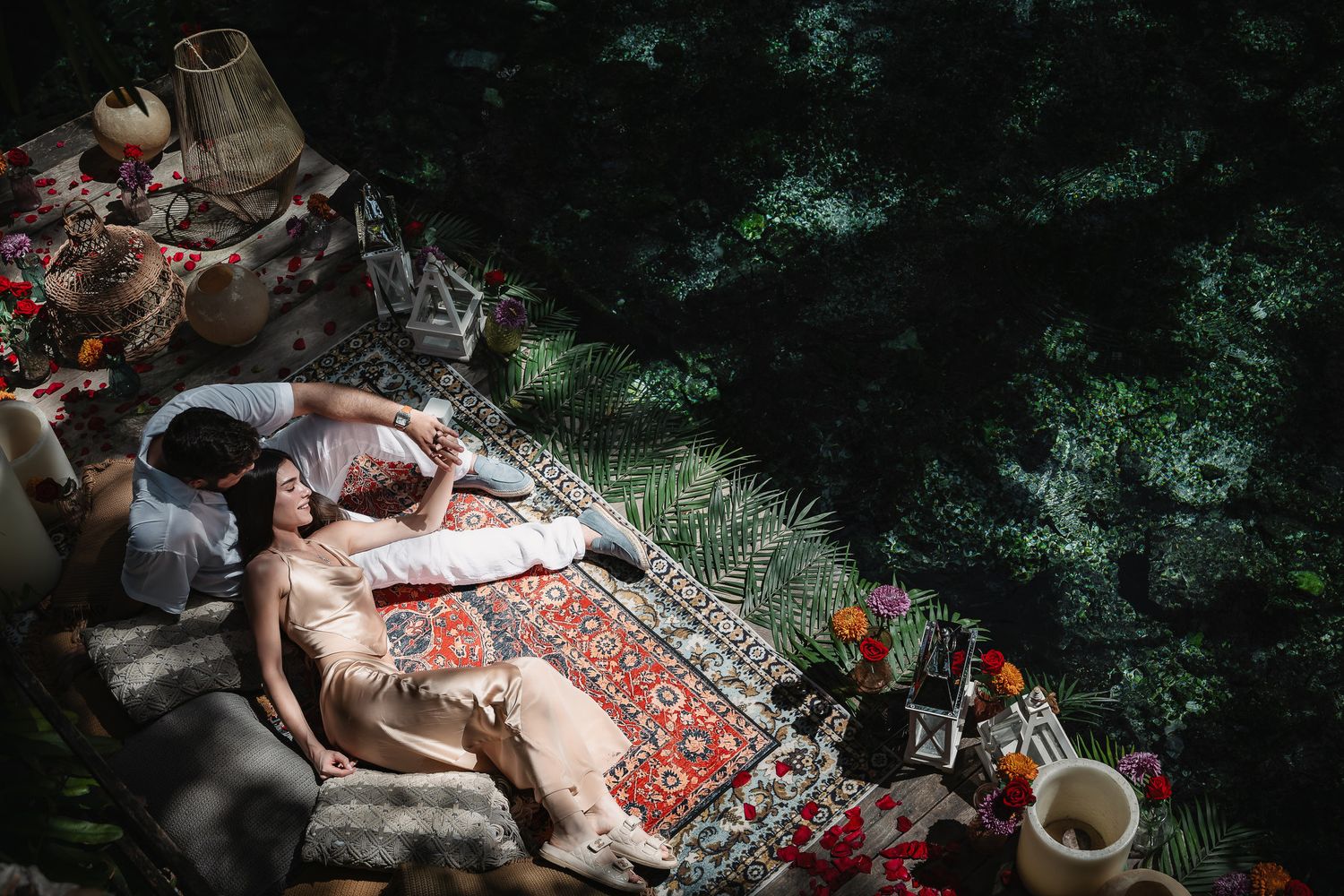 Couple lying on ornate rugs surrounded by ceramic pots and flowers in a lush garden setting.