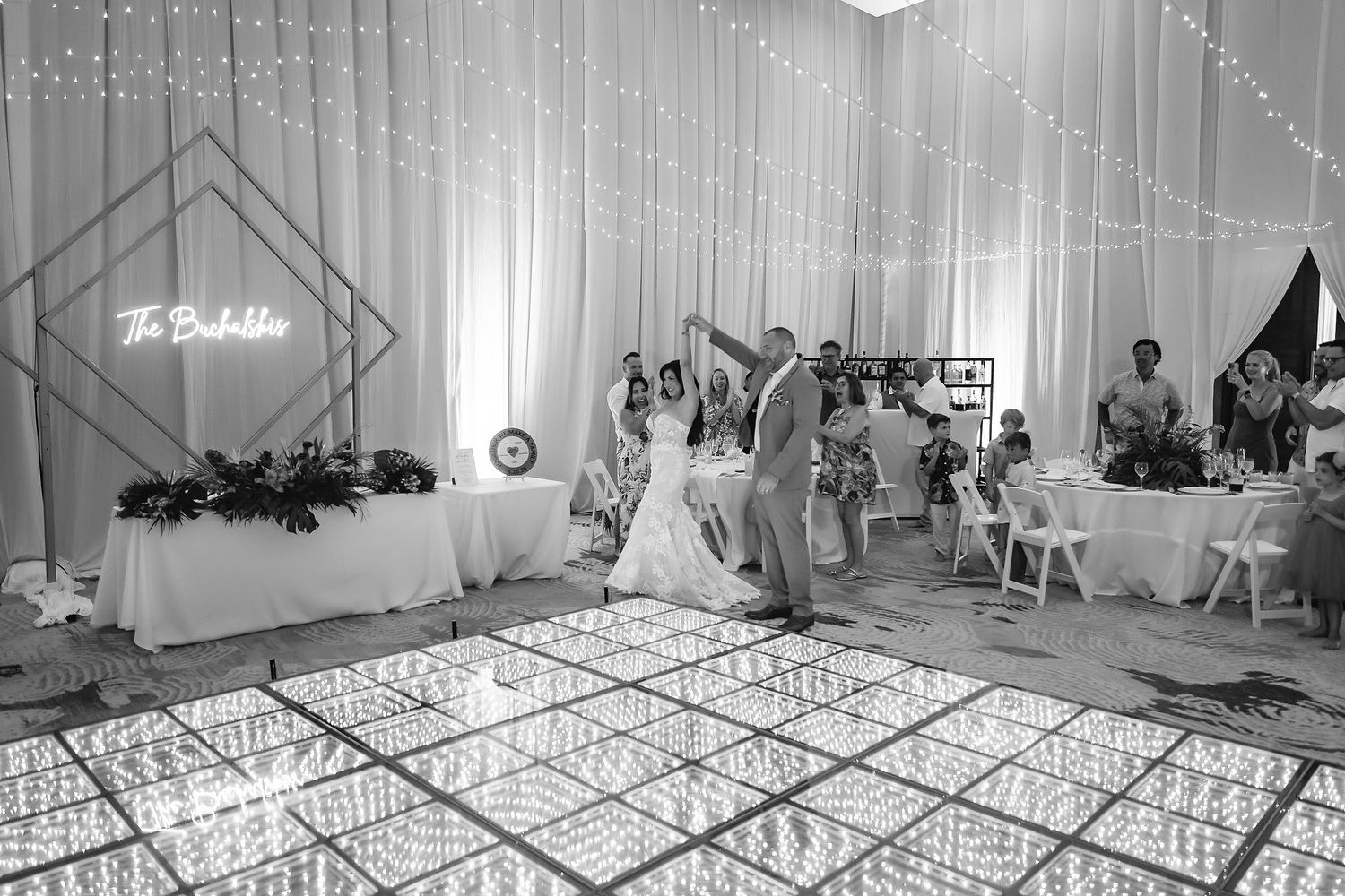 Couple shares first dance on illuminated floor at elegant wedding reception with fairy lights and guests seated nearby.