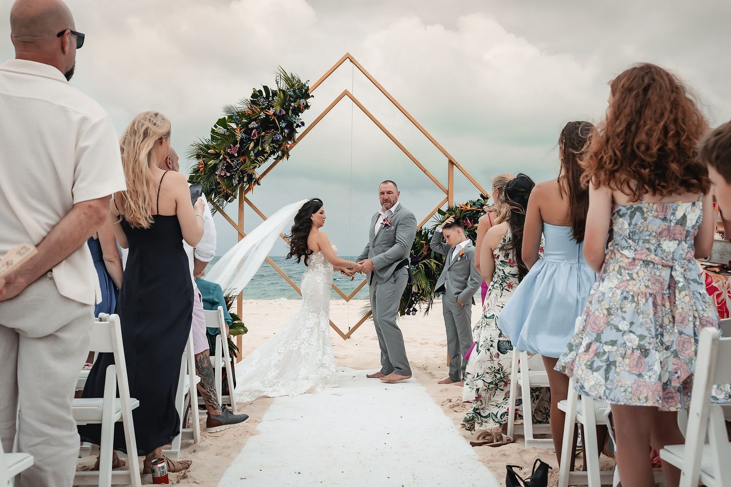 Bride and groom hold hands at a romantic beach wedding ceremony with a geometric floral arch backdrop.