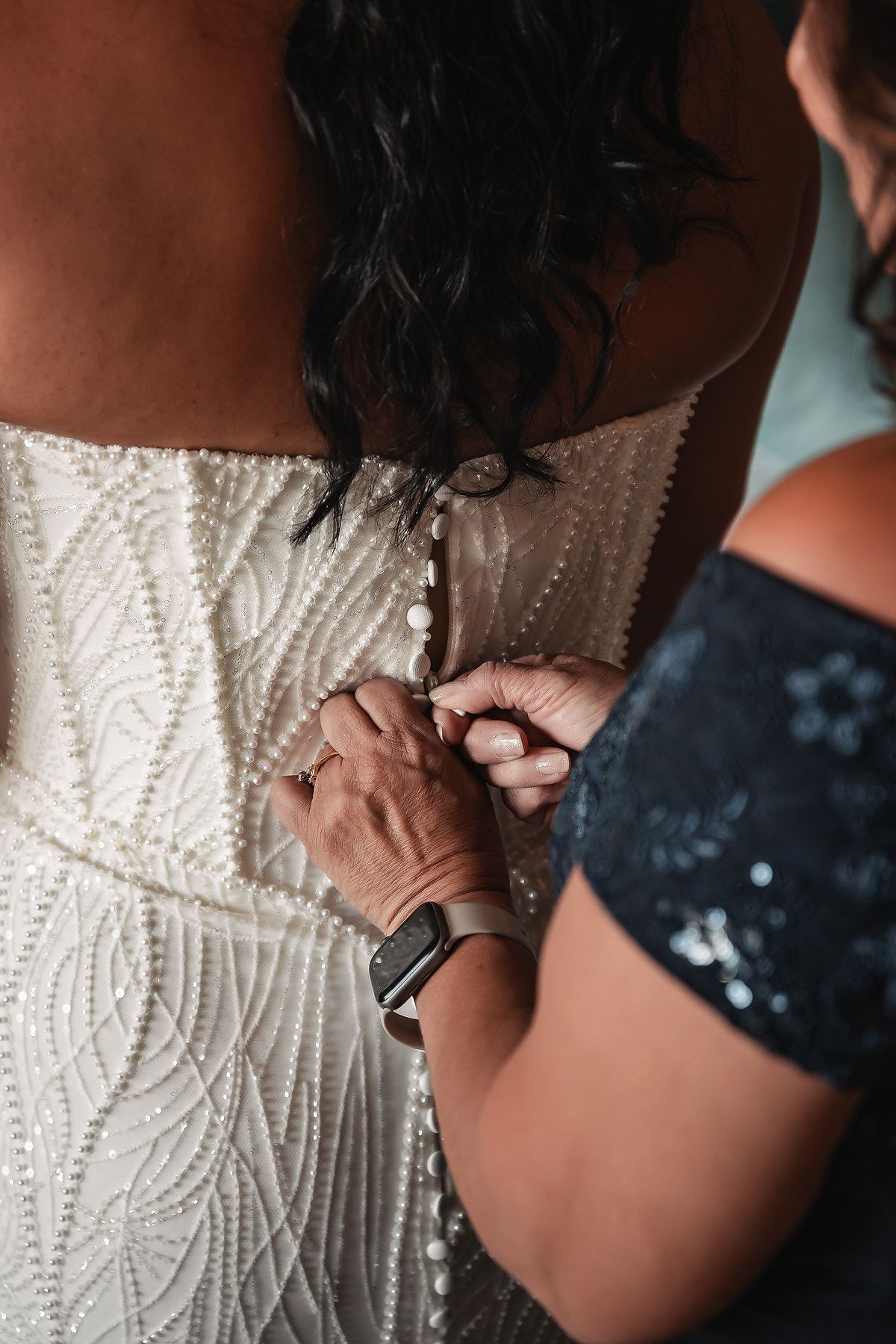 Helper buttoning the back of a beaded white wedding gown on a dark-haired bride during bridal preparations.