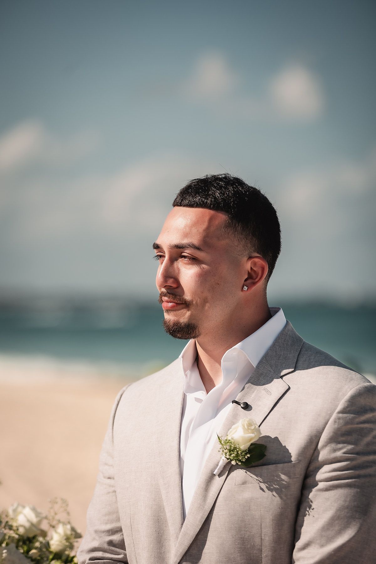 Groom in gray suit with white rose boutonniere standing on beach, gazing thoughtfully into distance on wedding day.