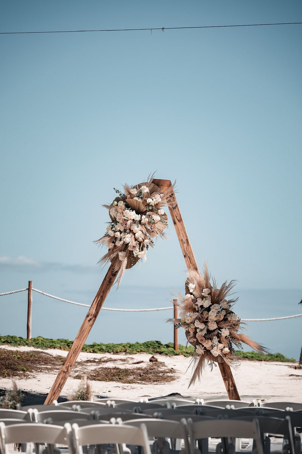Rustic wooden arch with floral arrangements on a sandy beach, set up for an outdoor wedding ceremony under a clear blue sky.
