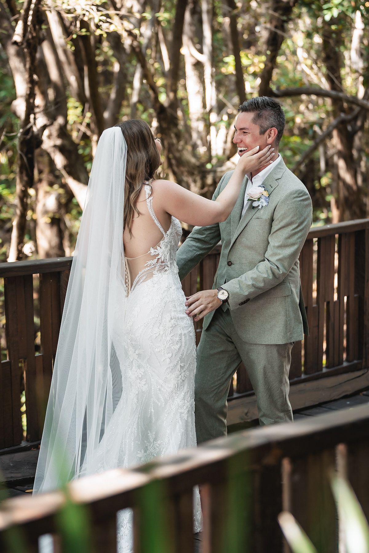 Bride in lace backless gown and veil tenderly touches grooms face on wooden deck surrounded by lush greenery.