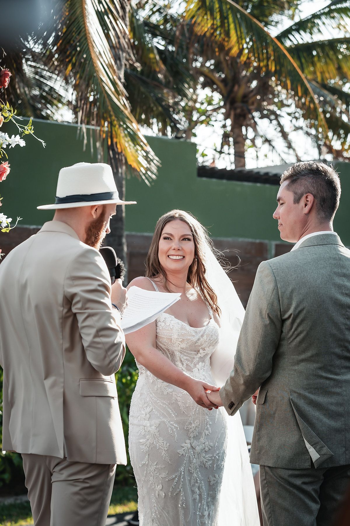 Bride and groom hold hands during outdoor wedding ceremony, officiant in white hat reads vows, palm trees in background.