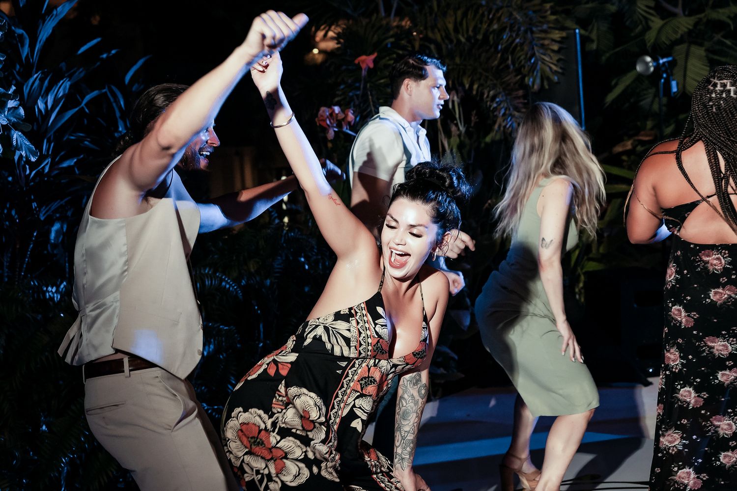 Group of people dancing energetically at an outdoor evening party, woman in floral dress raising arms joyfully.
