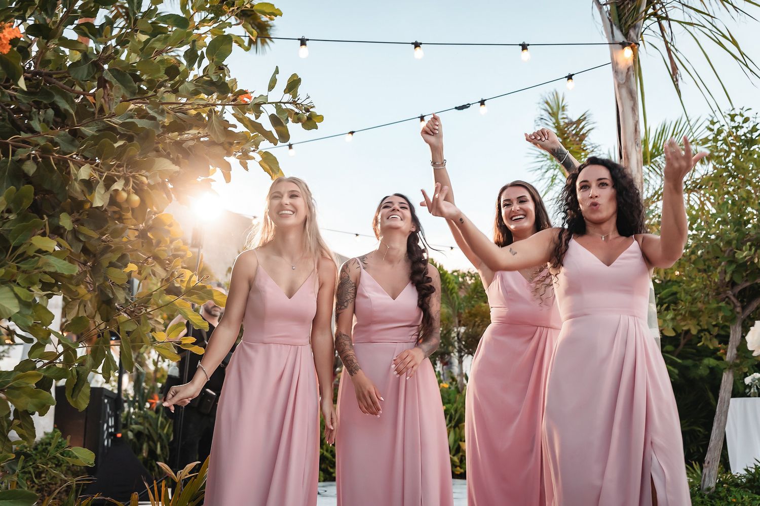 Four bridesmaids in pink satin gowns laugh and celebrate outdoors at a garden wedding reception with string lights.