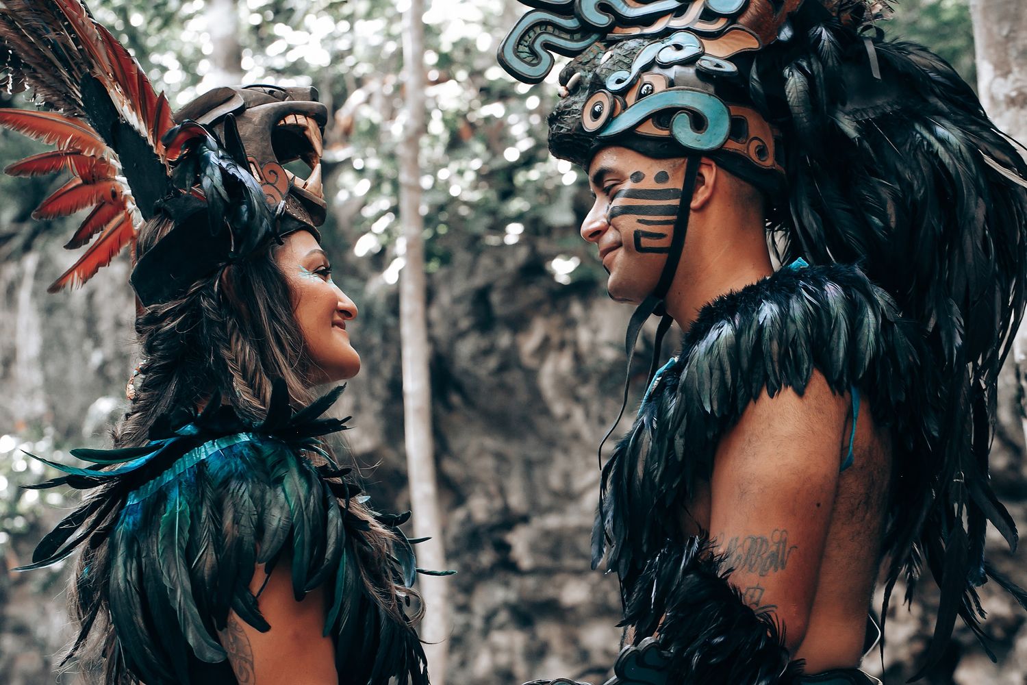 Two people in dark feathered Mayan ceremonial costumes face each other in a forest, with face paint and headdresses.