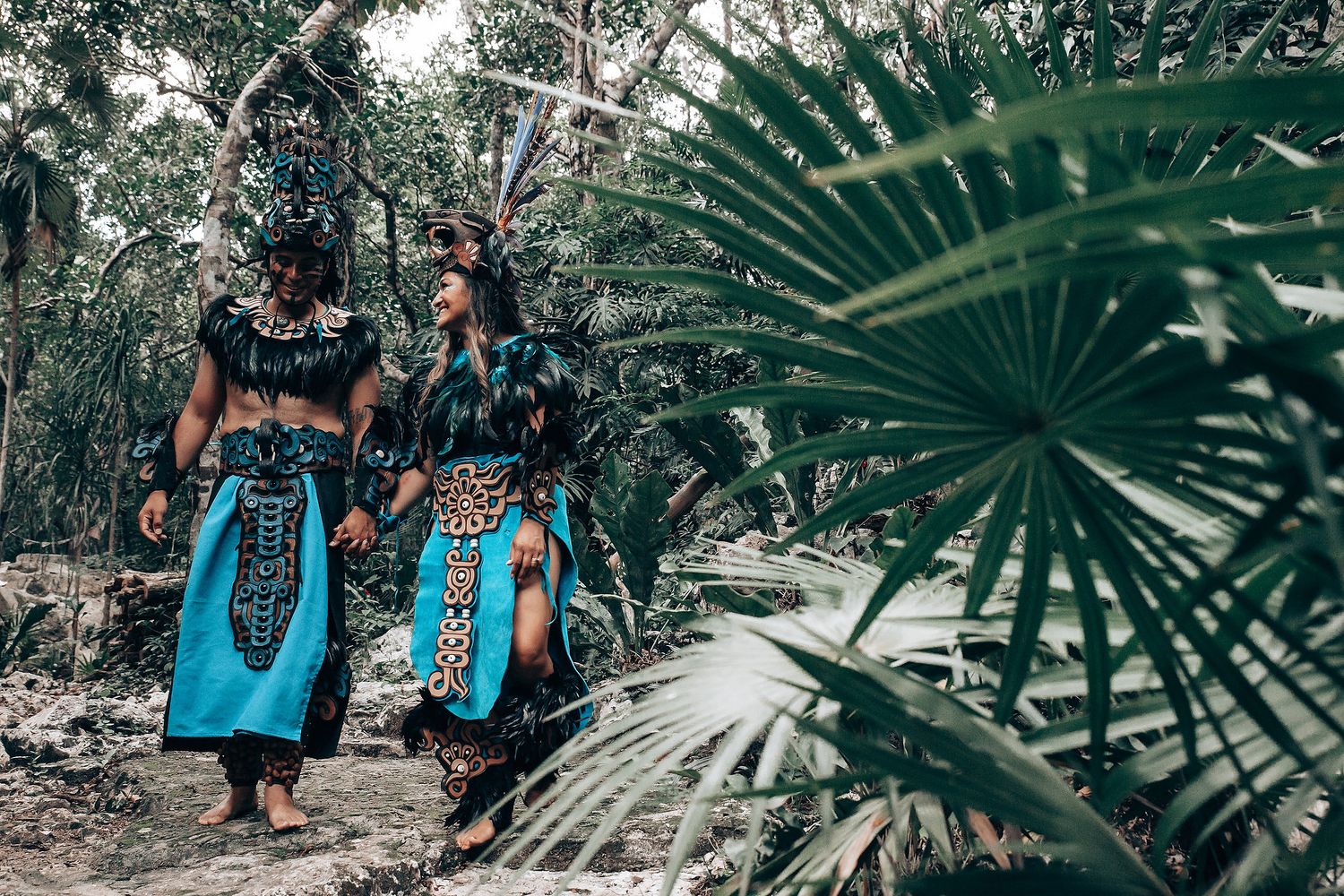 Couple in matching turquoise and black Mayan ceremonial costumes walking hand in hand through jungle path.