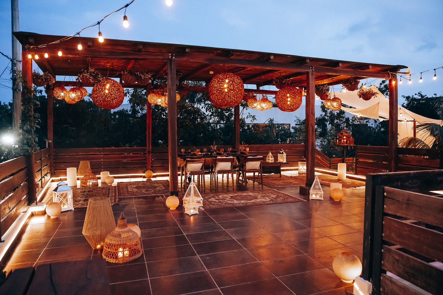 Cozy outdoor terrace at dusk with glowing rattan pendant lights, string lights, and decorative lanterns.