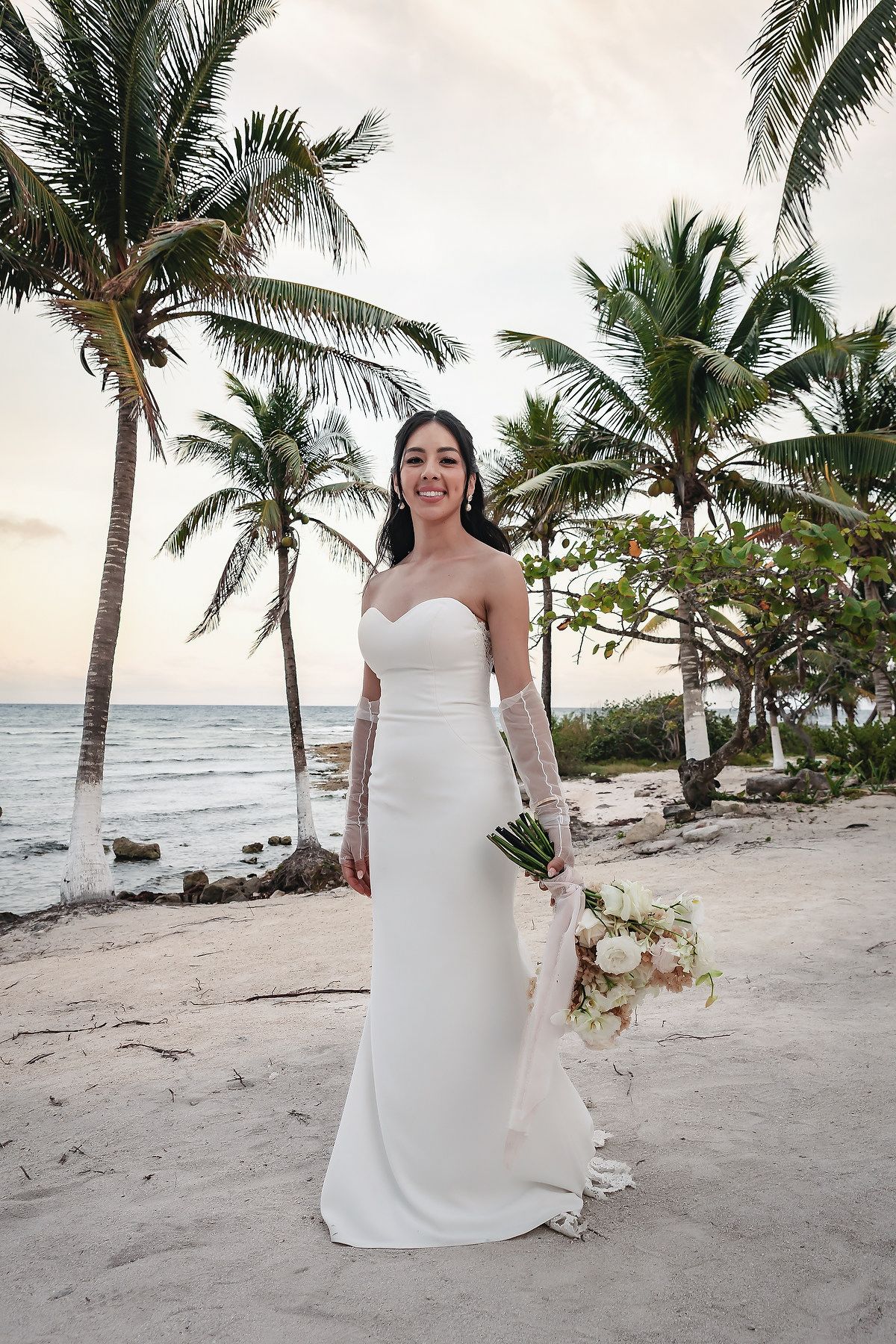 Bride in strapless white gown with lace gloves holds bouquet on tropical beach with palm trees at sunset.