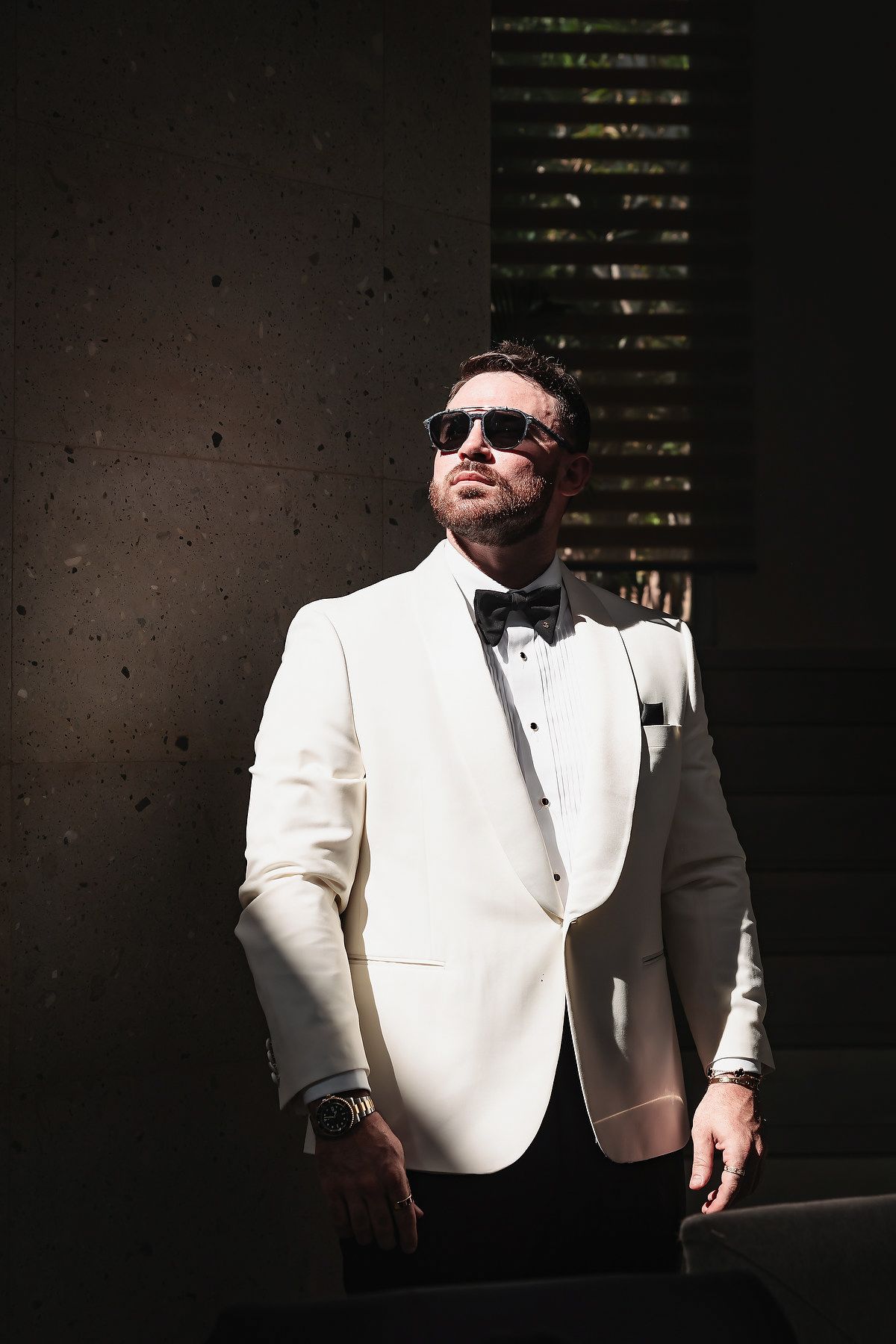 Man in white tuxedo jacket and bow tie wearing sunglasses poses confidently in dramatic lighting.