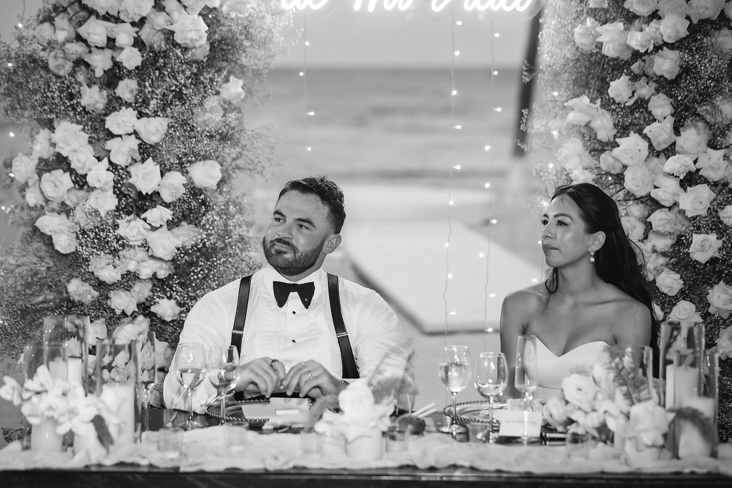 Bride and groom smiling at sweetheart table adorned with white floral arrangements at elegant wedding reception.