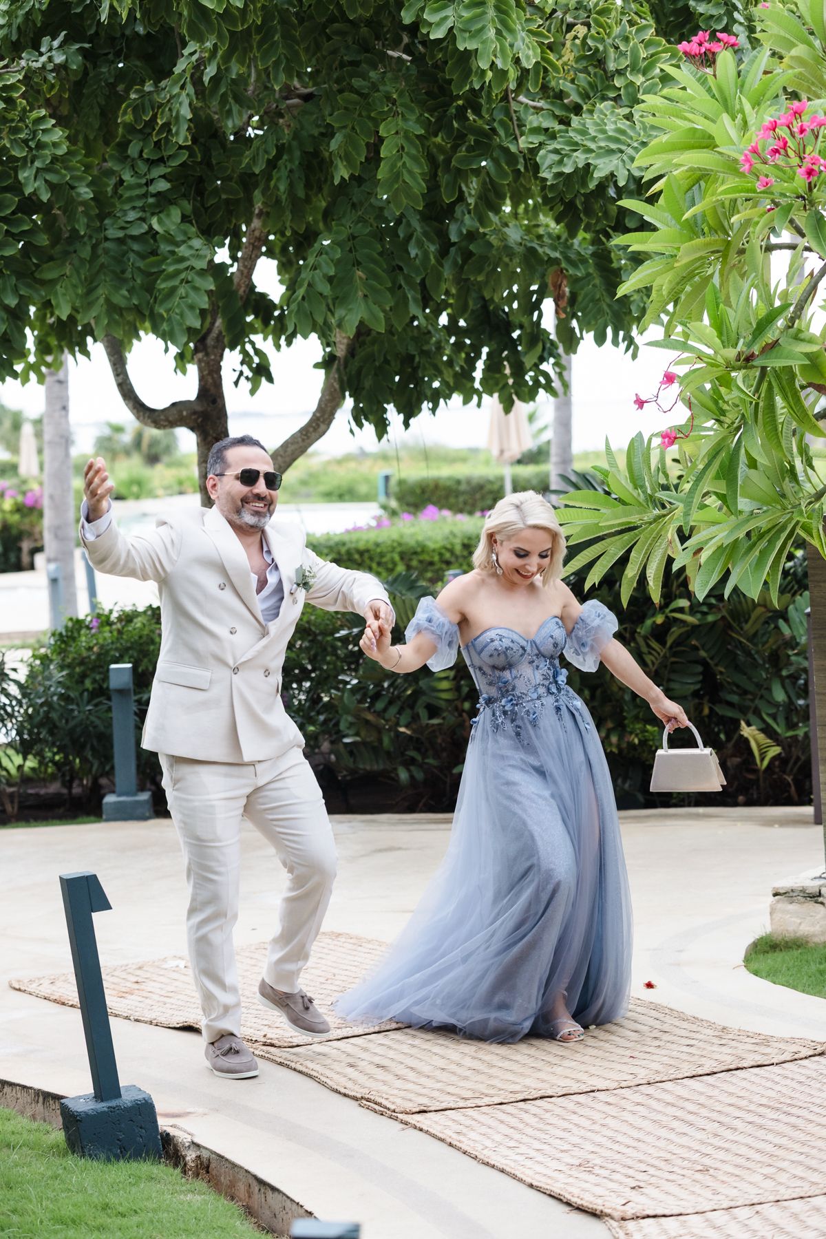 Elegant couple dancing outdoors, man in beige suit, woman in blue floral off-shoulder gown, tropical garden setting.