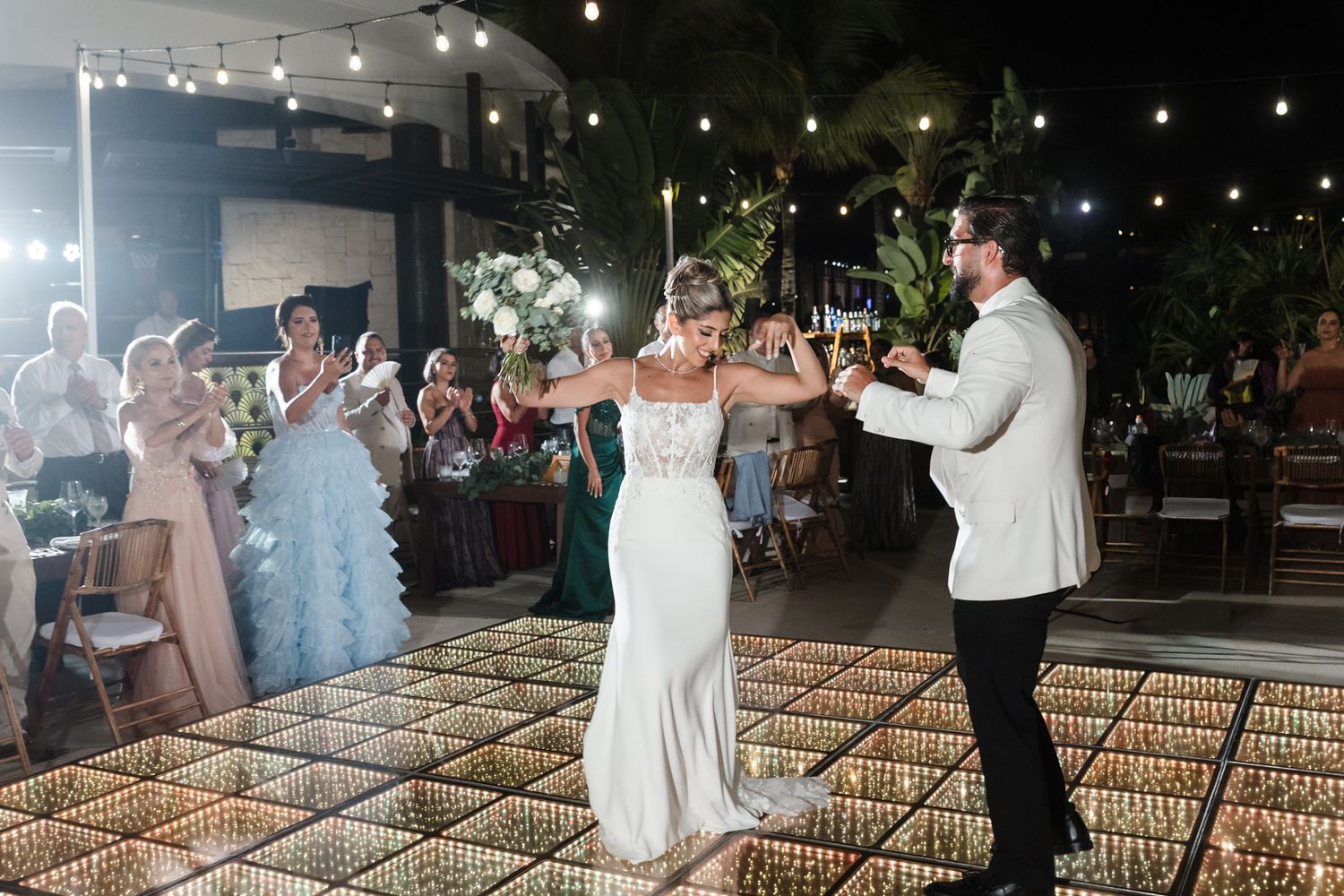 Bride and groom share first dance on illuminated floor at outdoor evening wedding reception with guests applauding.