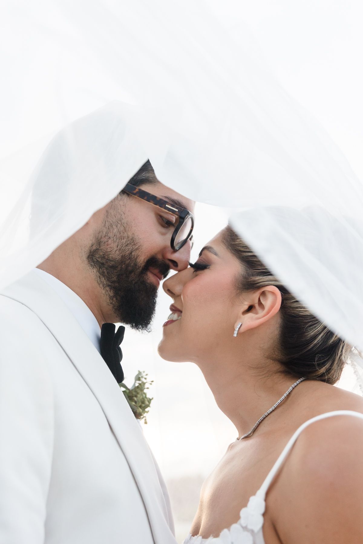 Romantic wedding couple about to kiss under a flowing veil, groom wearing glasses and white suit.