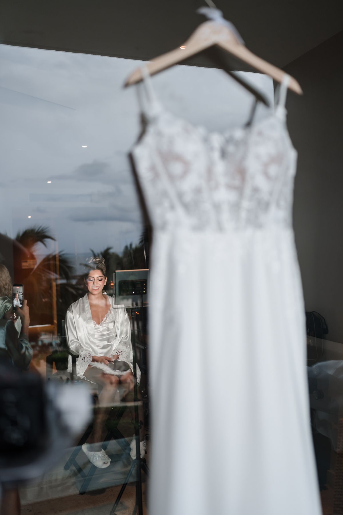 A white wedding dress hangs on a hanger as a bride in a satin robe sits in the background during bridal preparations.