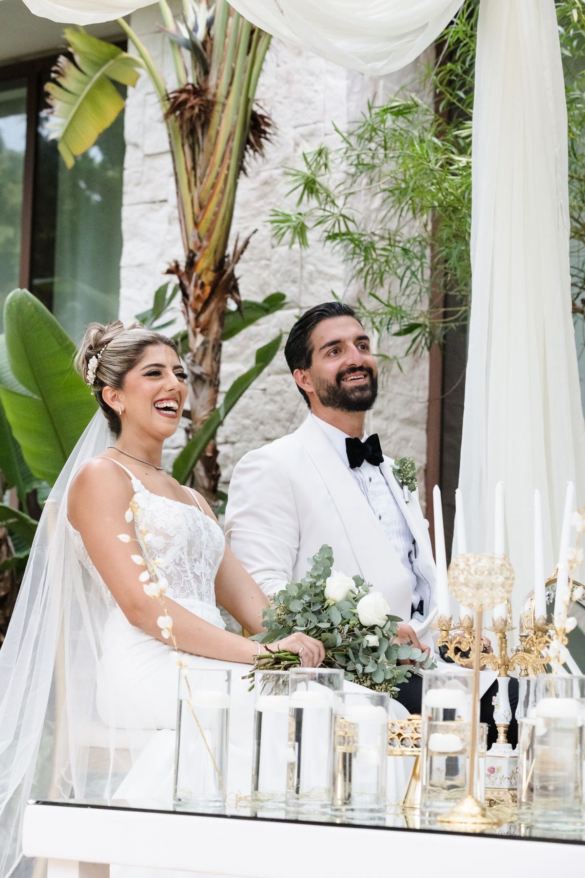 Smiling bride and groom in white attire stand at decorated wedding table with candles and florals outdoors.