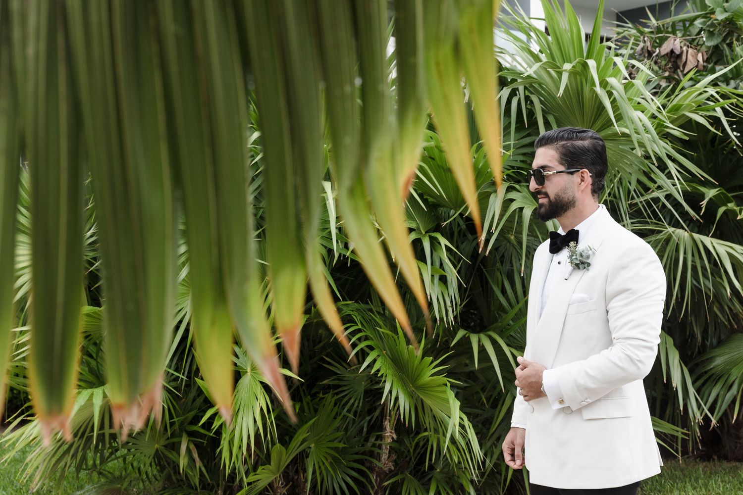 A dapper groom in a white tuxedo jacket and bow tie stands among lush tropical palm greenery outdoors.