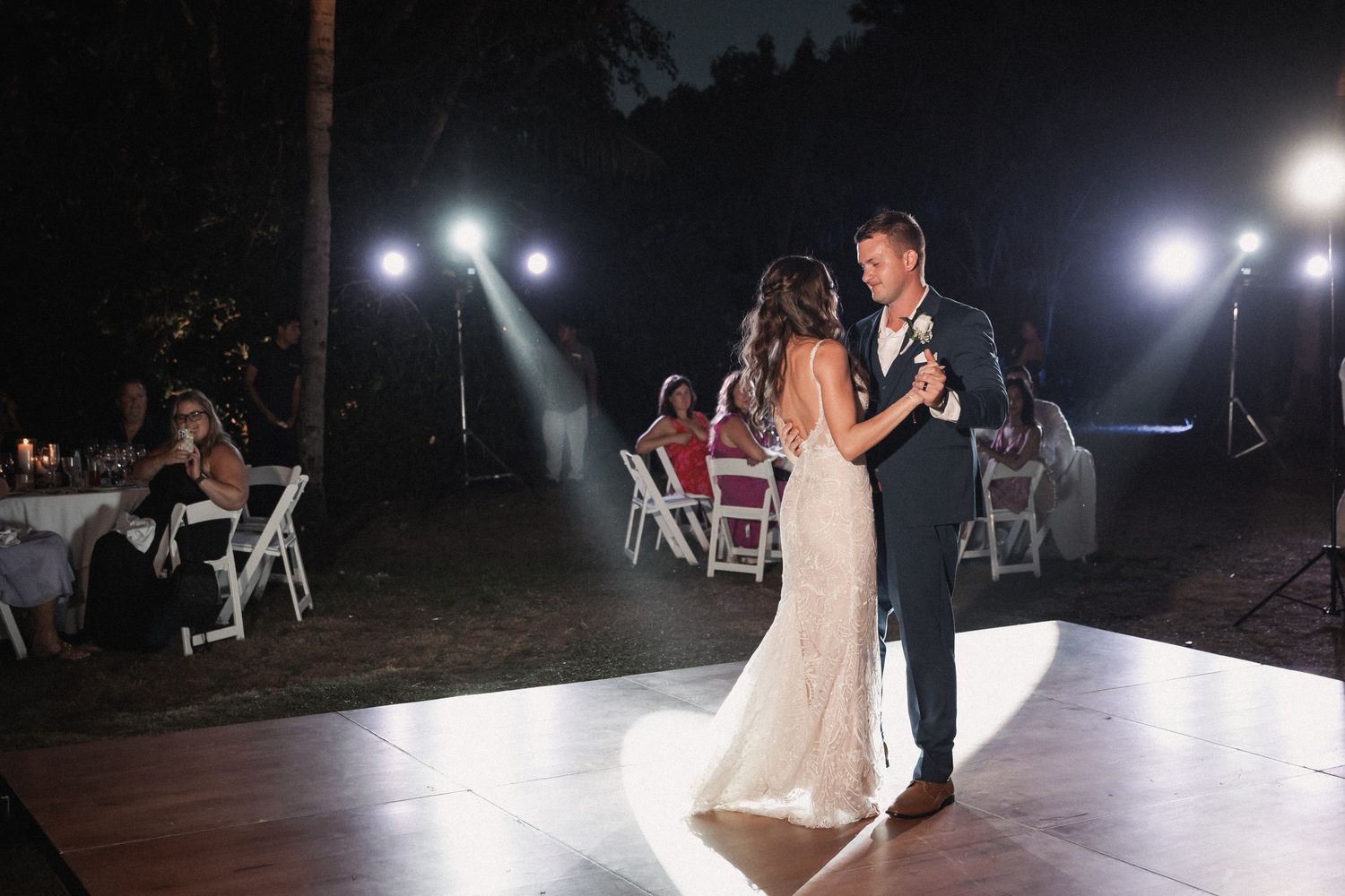 Bride and groom share first dance on white dance floor at romantic outdoor nighttime wedding reception.